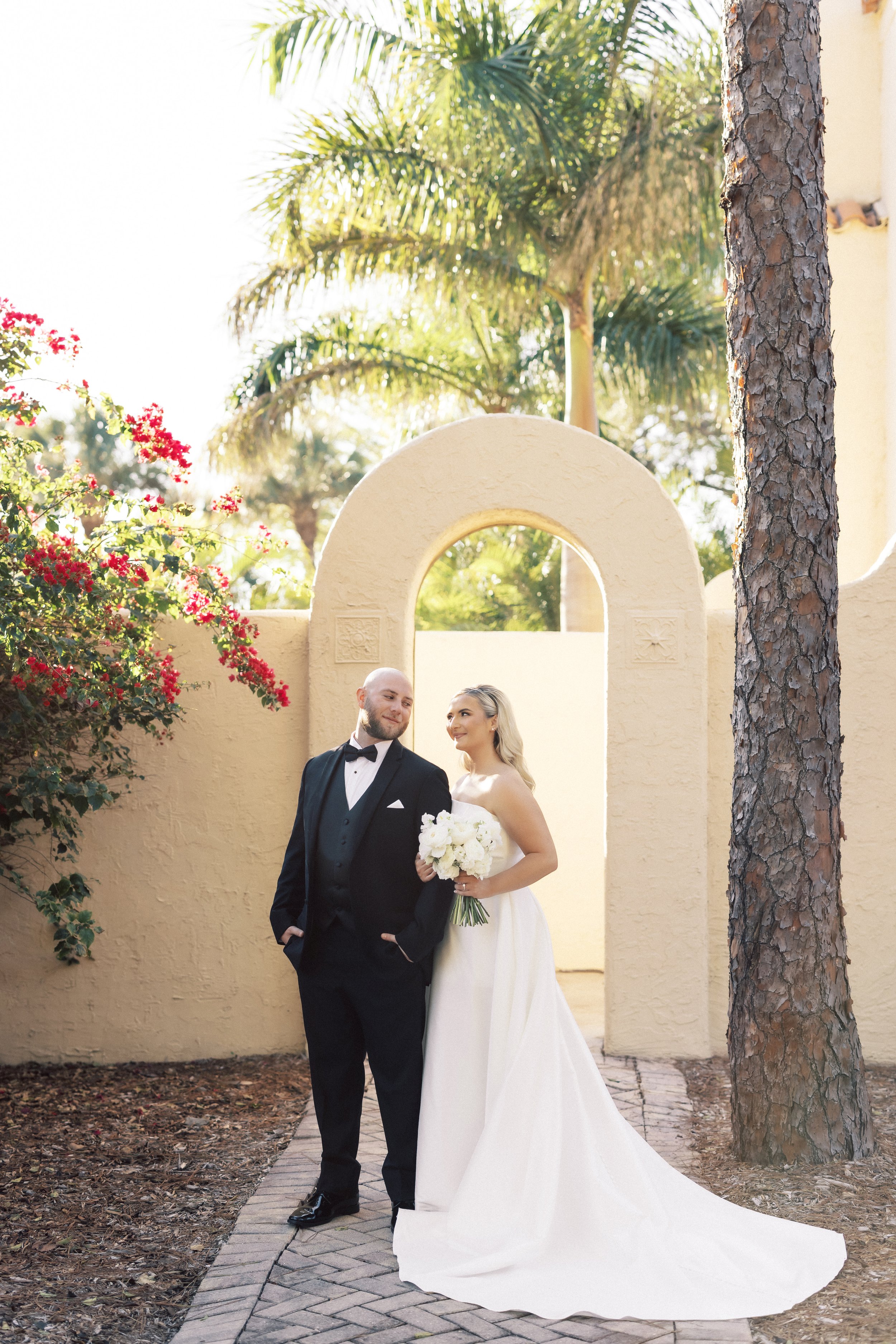 A bride and groom standing outdoors under an archway, the bride holding a bouquet of white flowers, trees and plants in the background.