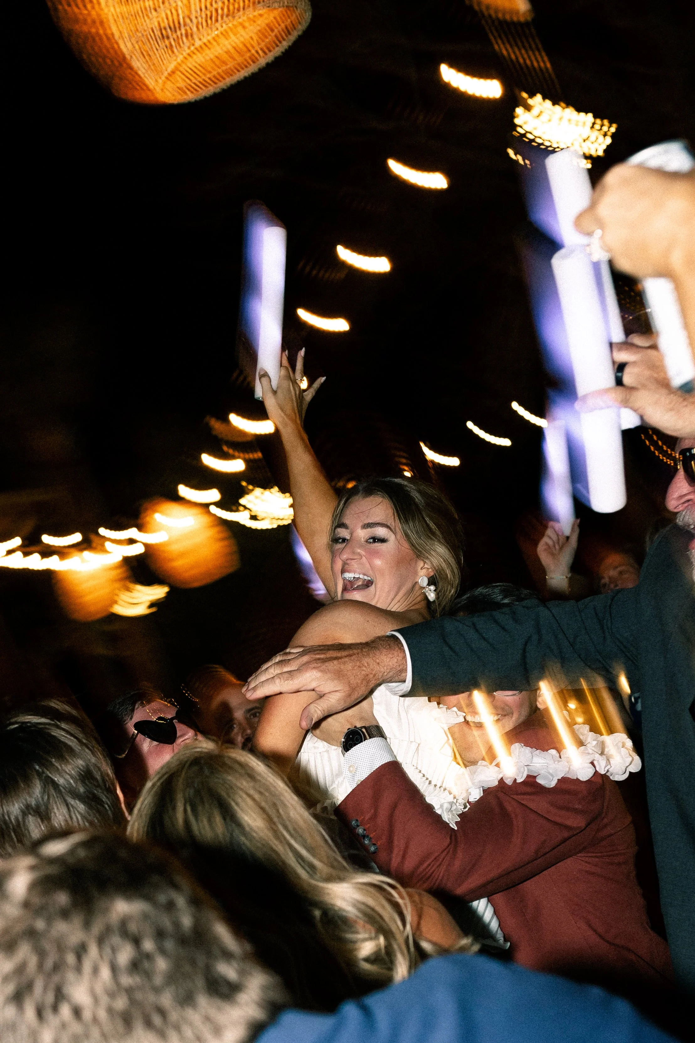 People celebrating, with a woman in a wedding dress smiling amid a lively crowd, holding up a glowing light stick.