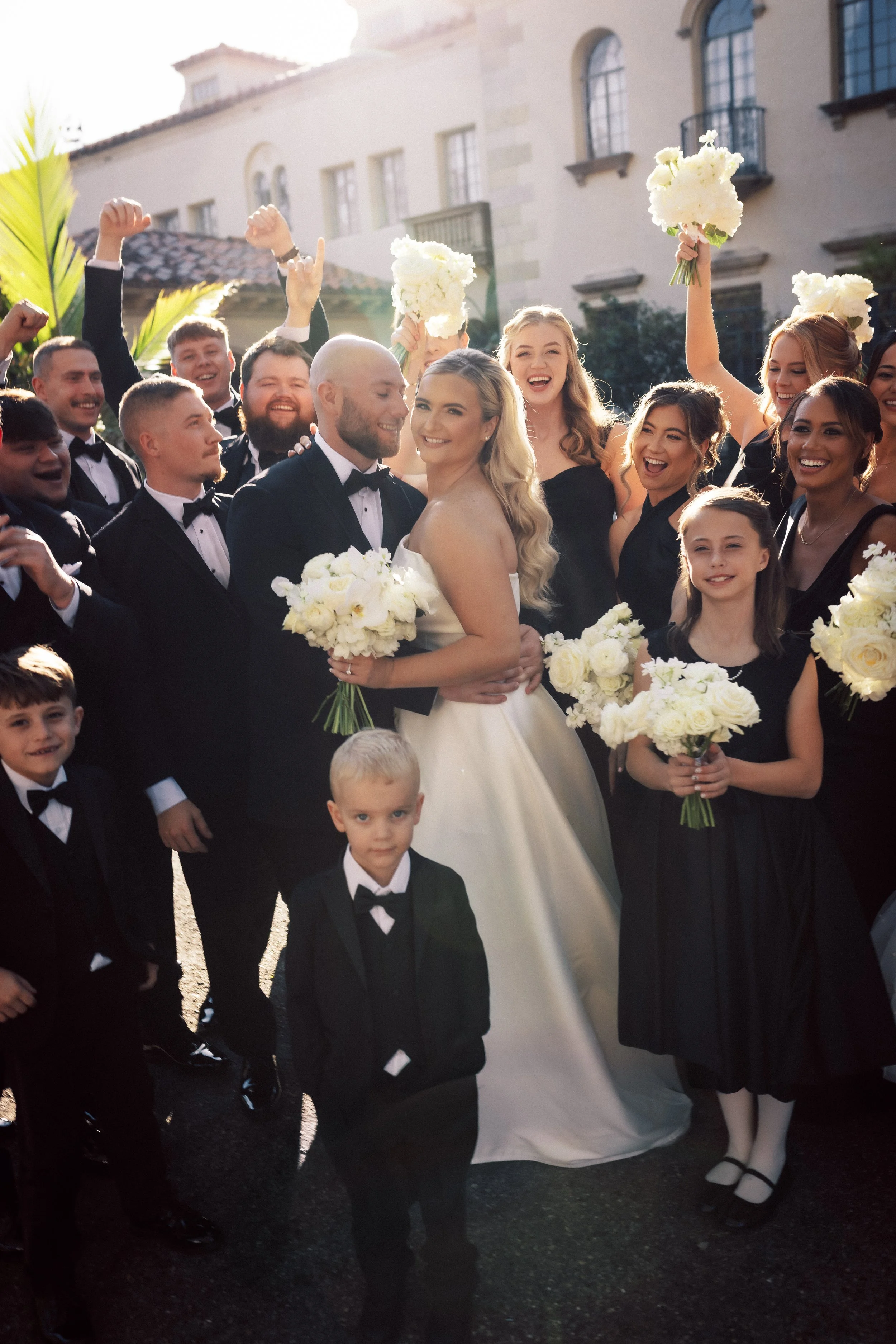 A wedding party outdoors with smiling bride and groom in the center, surrounded by bridesmaids, groomsmen, and children all dressed in formal attire, holding white bouquets and celebrating.