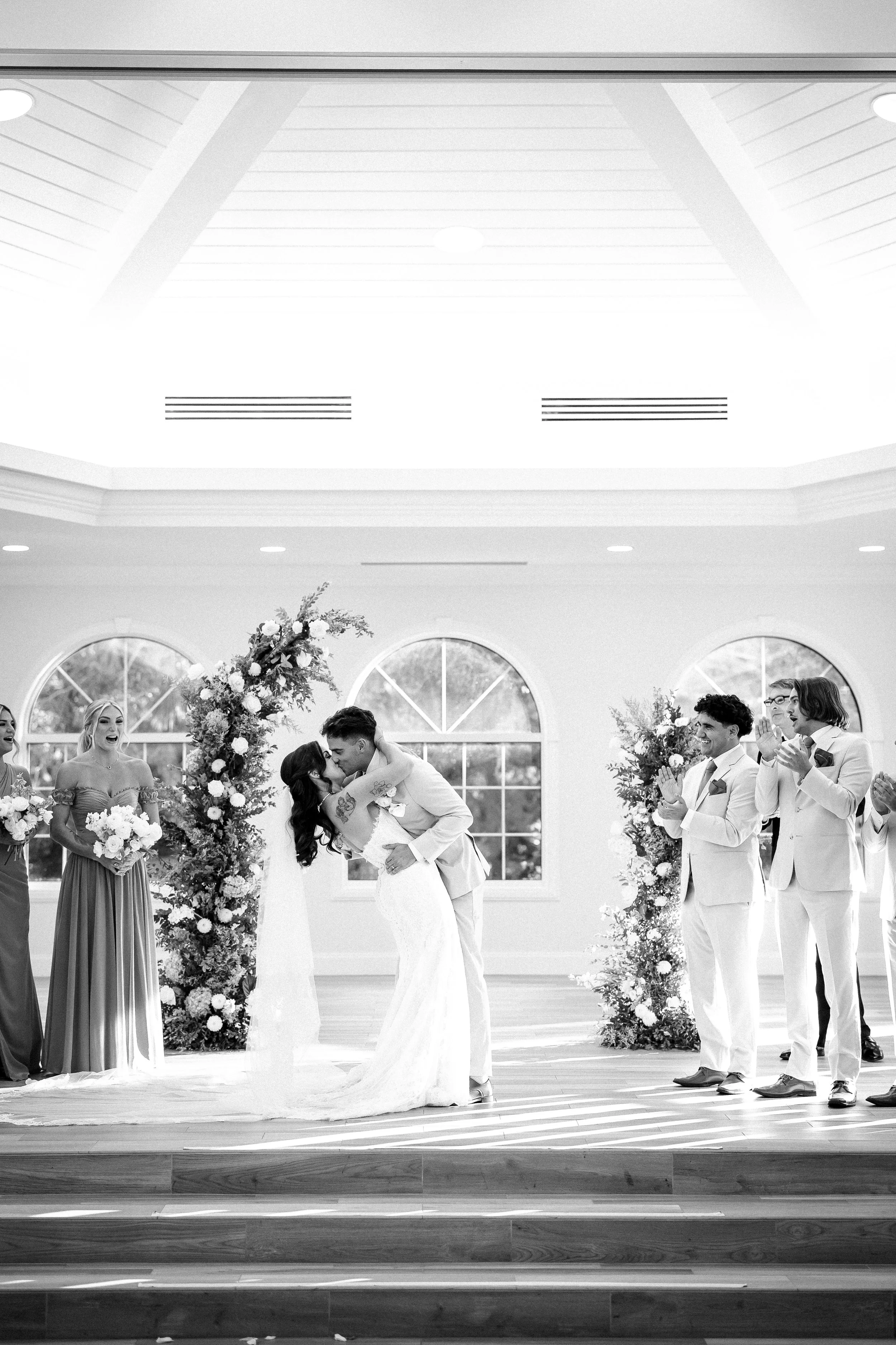 Black and white wedding ceremony with a couple kissing at the altar, surrounded by bridesmaids and groomsmen clapping and smiling, in a bright room with large arched windows and floral decorations.