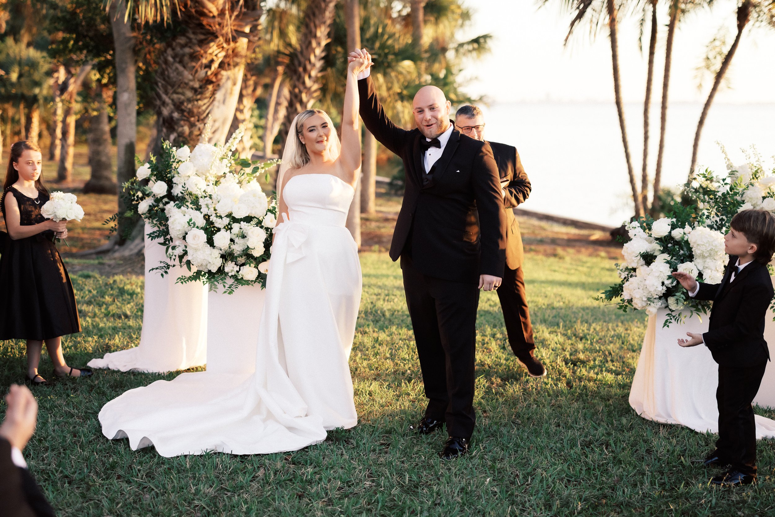 Bride and groom celebrating their wedding outdoors near a lake during sunset, with flower arrangements and children dressed in formal attire.