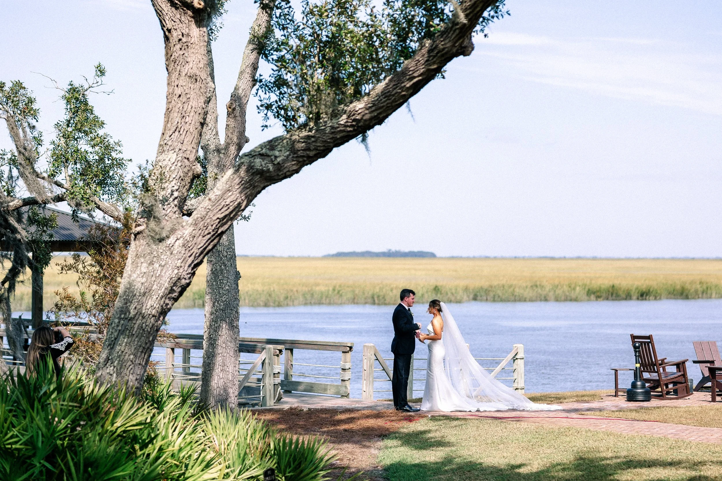 A bride and groom standing on a wooden deck by a body of water, holding hands, during their wedding ceremony outdoors with trees and grass nearby.