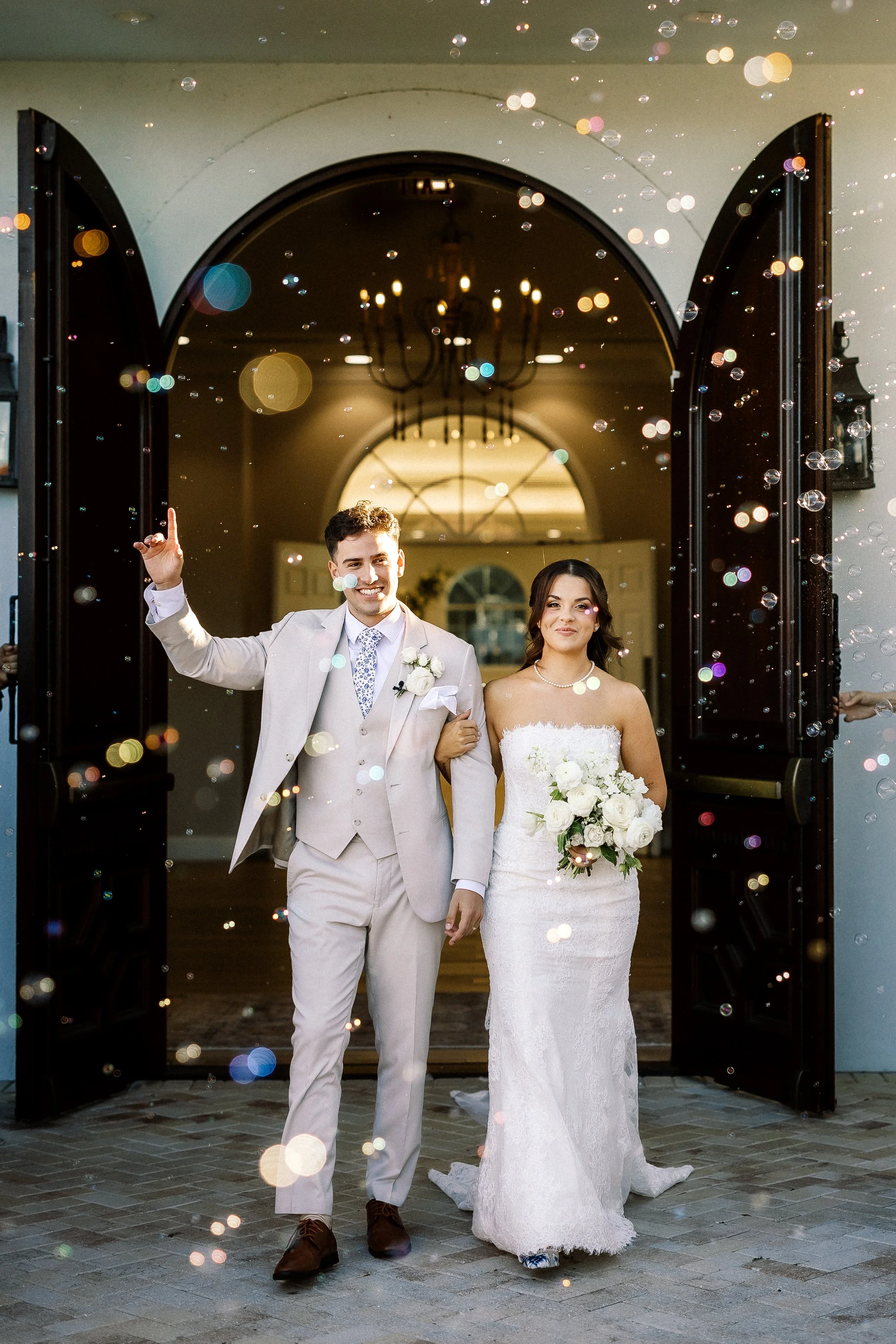 A newlywed couple walking out of a building through open doors, celebrating their wedding, with bubbles floating around them.
