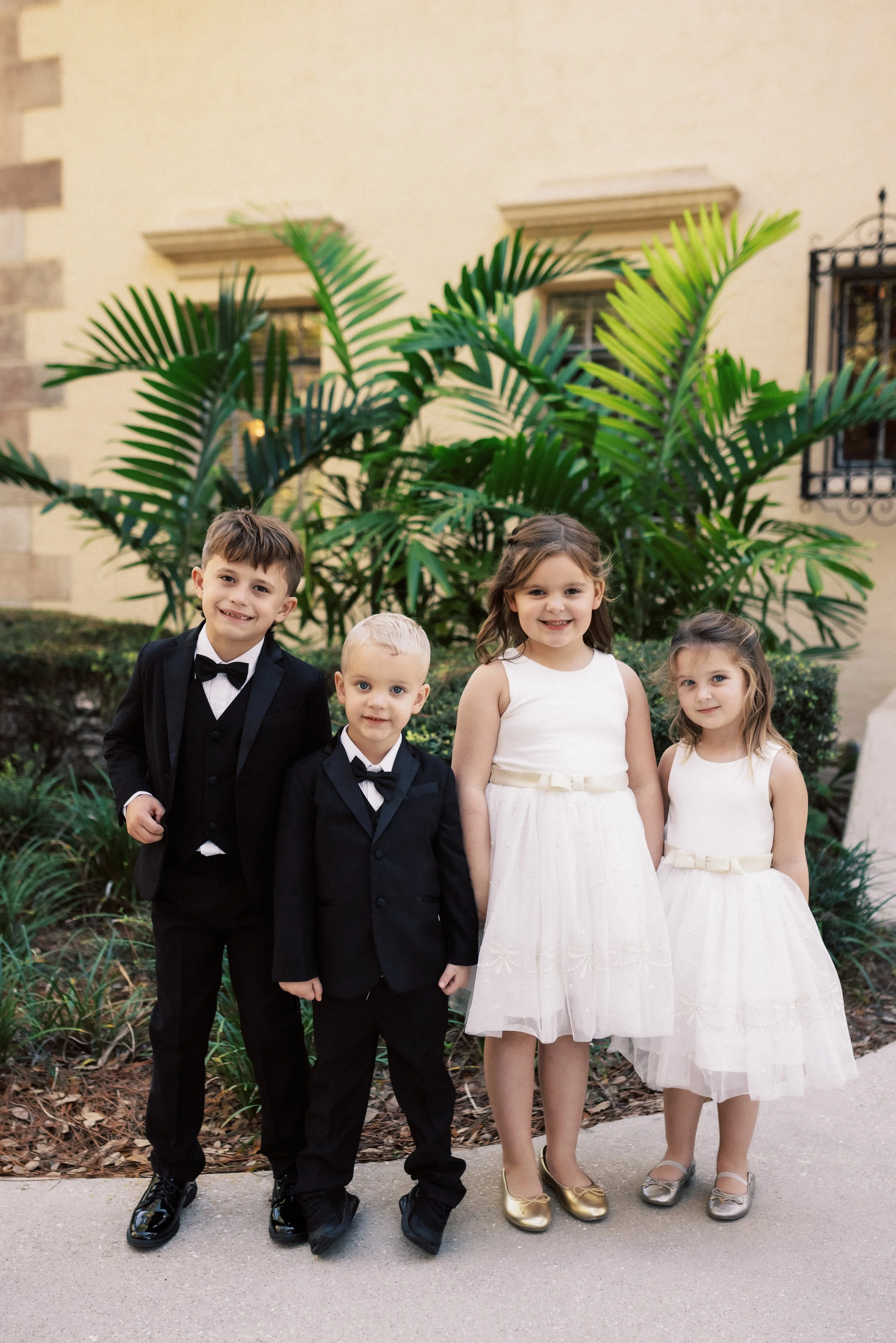 Four children dressed in formal attire standing outdoors in front of large green plants. Two boys in black suits and bow ties, two girls in white dresses with cream bows.