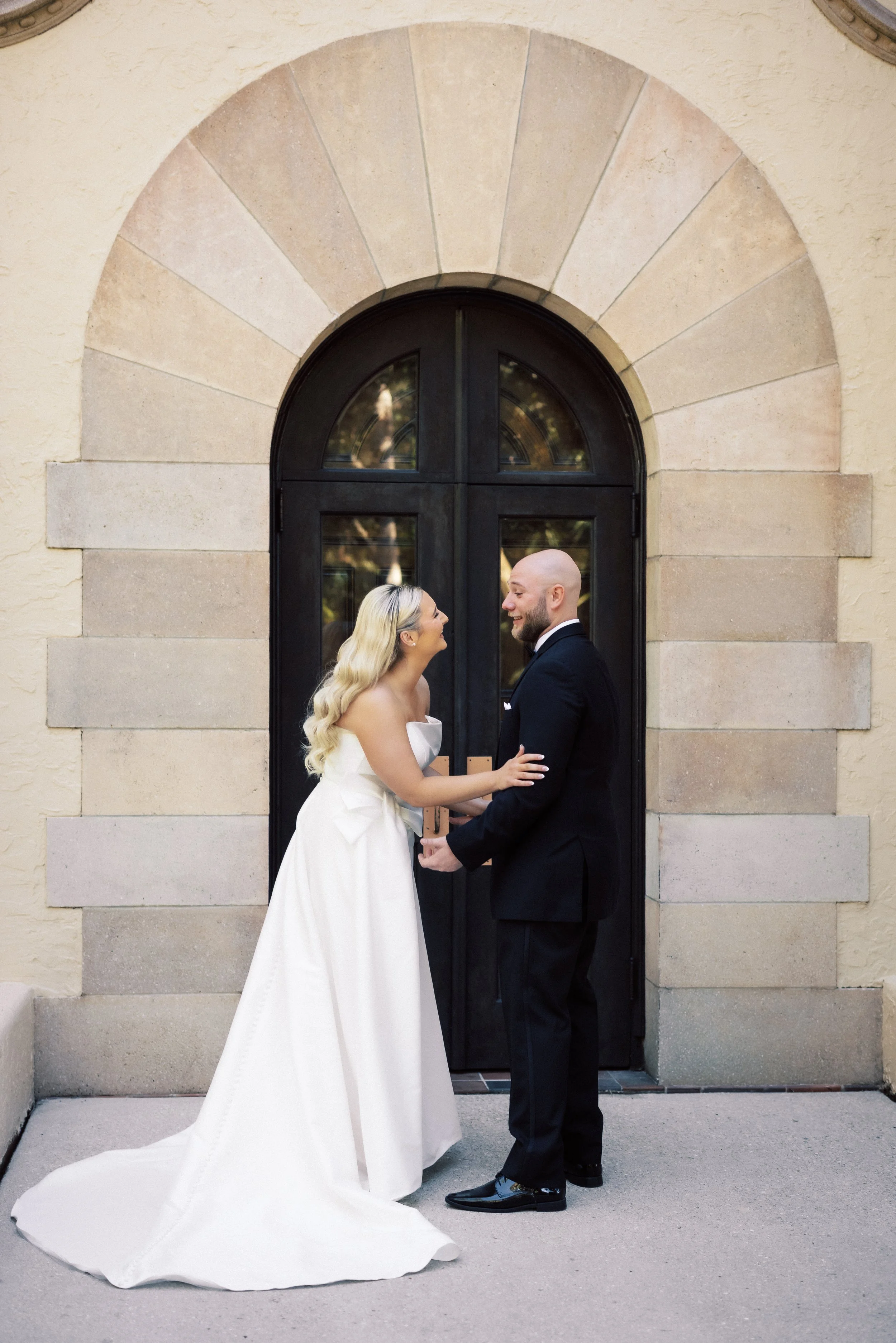 Bride and groom standing face to face in front of a black arched door, smiling. The bride is wearing a white wedding gown and the groom is in a black suit. They are holding hands.