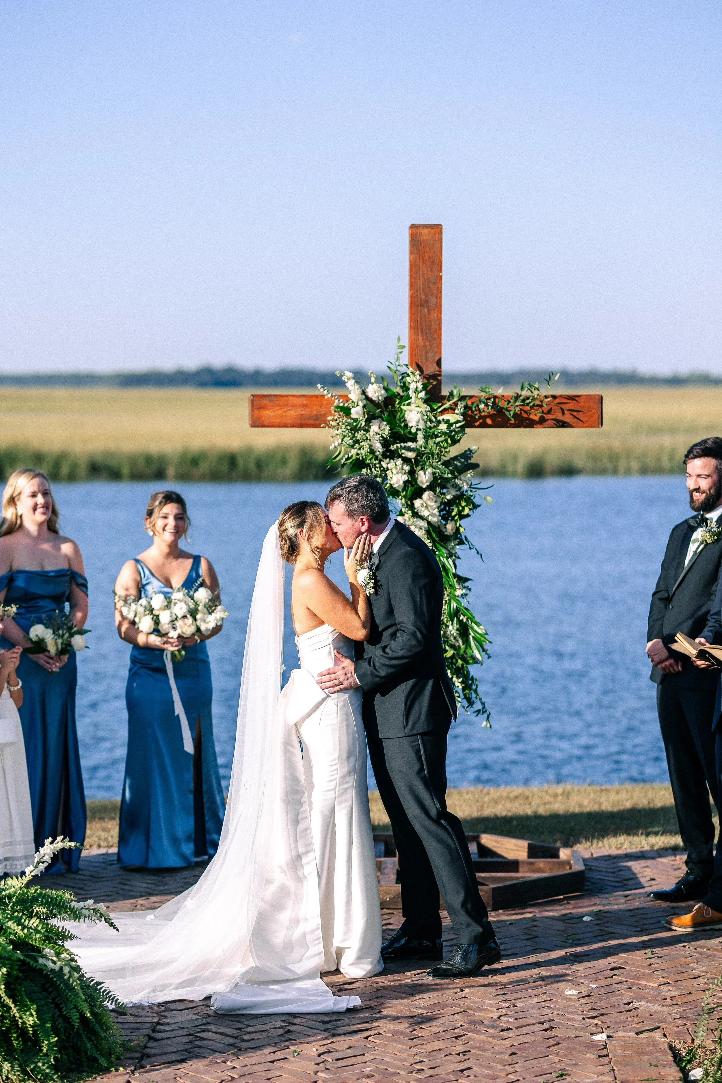 A wedding ceremony by a lake featuring a couple kissing under a wooden cross decorated with white flowers and greenery, with bridesmaids and groomsmen standing nearby on a brick path.