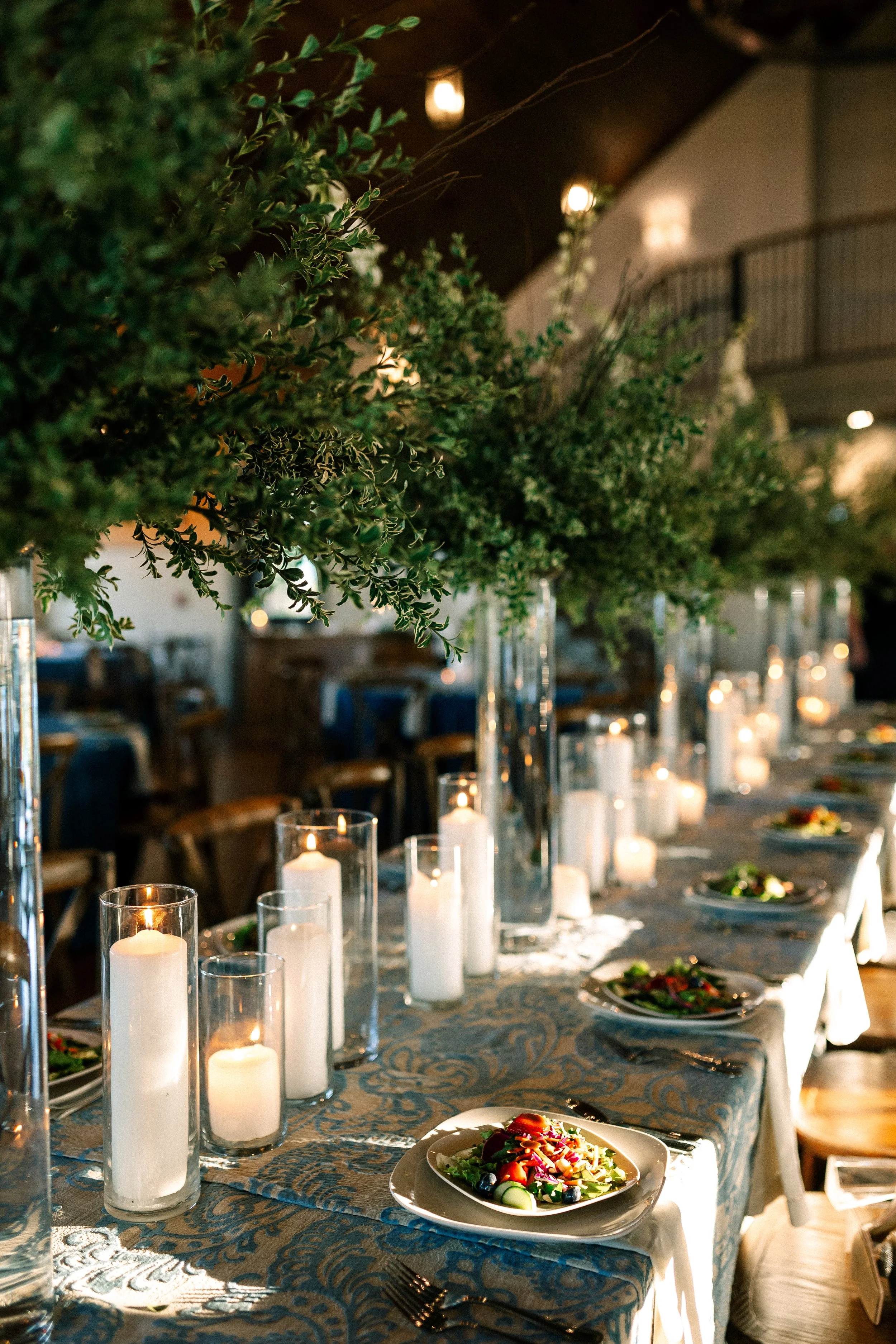 Decorated dining table with plates of salad, surrounded by lit candles in glass holders, greenery centerpieces, and set in an elegant restaurant with hanging lights and a balcony.