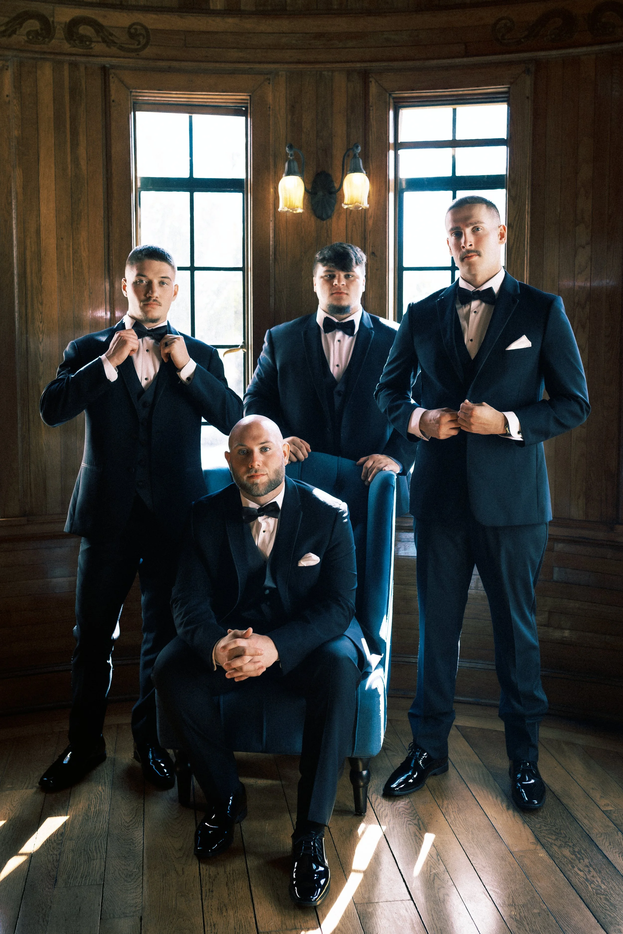 Five men dressed in formal tuxedos with bow ties inside a wood-paneled room with large windows and vintage lighting, posing for a photo.