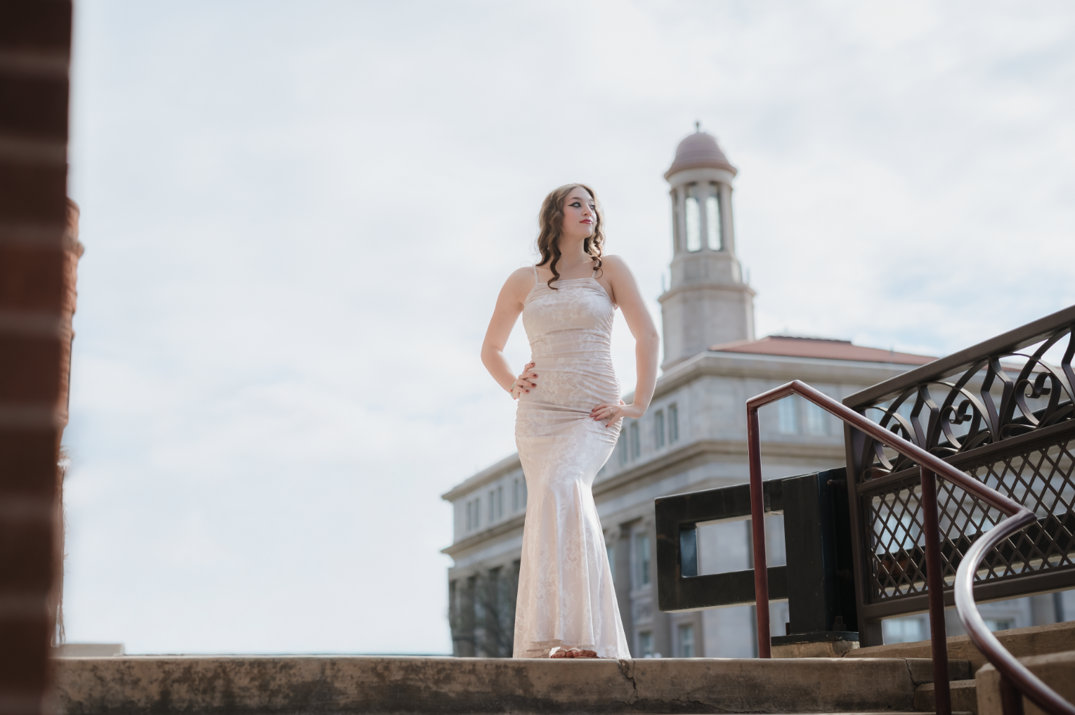 A woman in an elegant, form-fitting, sleeveless white dress stands on outdoor stairs next to a railing, with a historic building and clock tower in the background on a partly cloudy day.