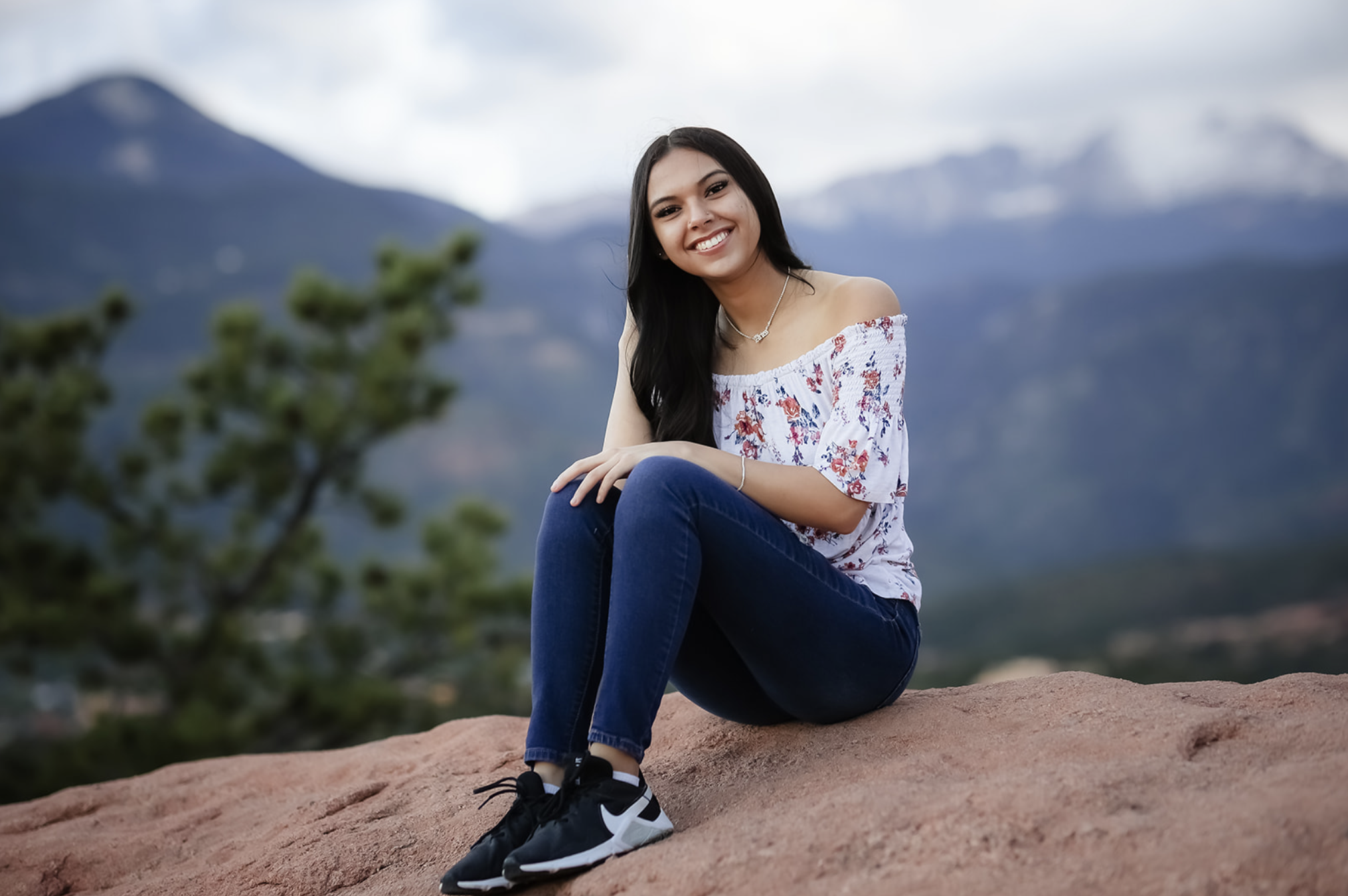 A young woman with long dark hair smiling at the camera, sitting on a rock in a mountainous outdoor setting with blurred mountains and sky in the background.