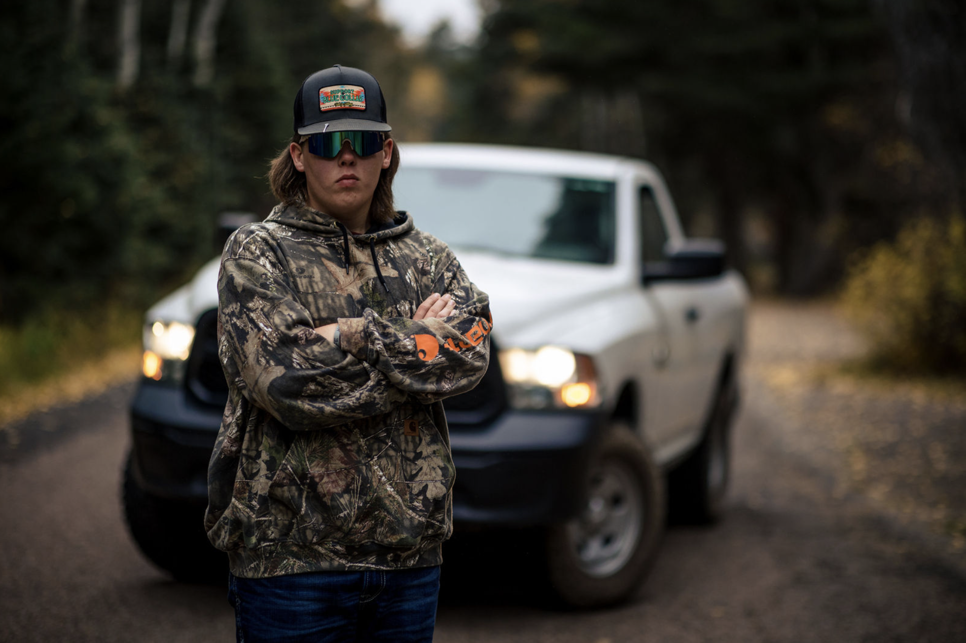 A young man with long hair, wearing a camouflage hoodie, cross-armed, standing in front of a white SUV with its headlights on, on a dirt road in a wooded area.