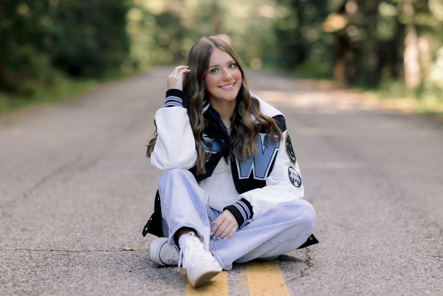 A young woman with long brown hair and blue eyes sitting cross-legged on a paved road in a wooded area, smiling at the camera. She is wearing a white and black varsity jacket with patches, a black shirt, light blue jeans, and white sneakers.