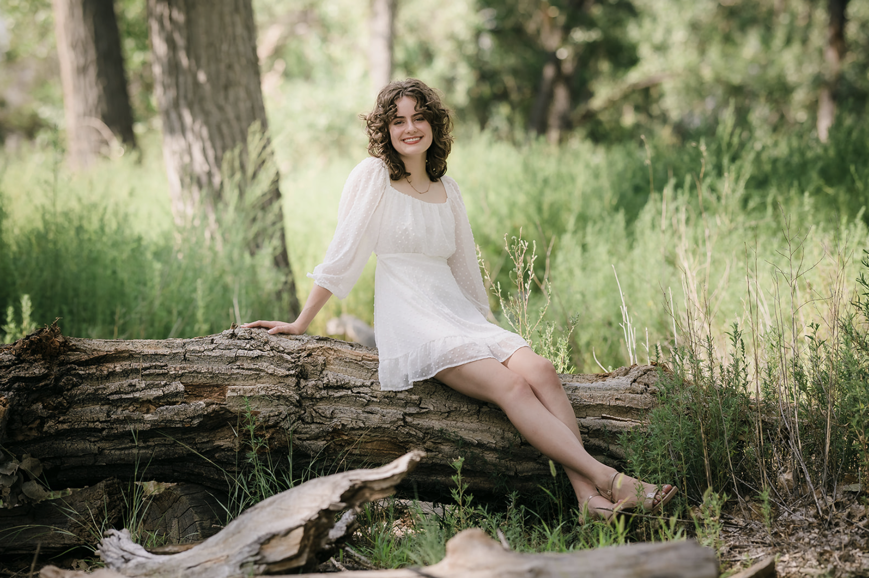 Woman in white dress sitting on a fallen tree in a forested area.