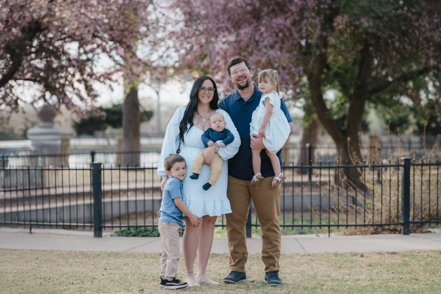 A young girl in a yellow dress with ruffled sleeves and black buttons sitting on a person's shoulder outdoors, smiling and looking at someone out of frame, with trees and greenery in the background.