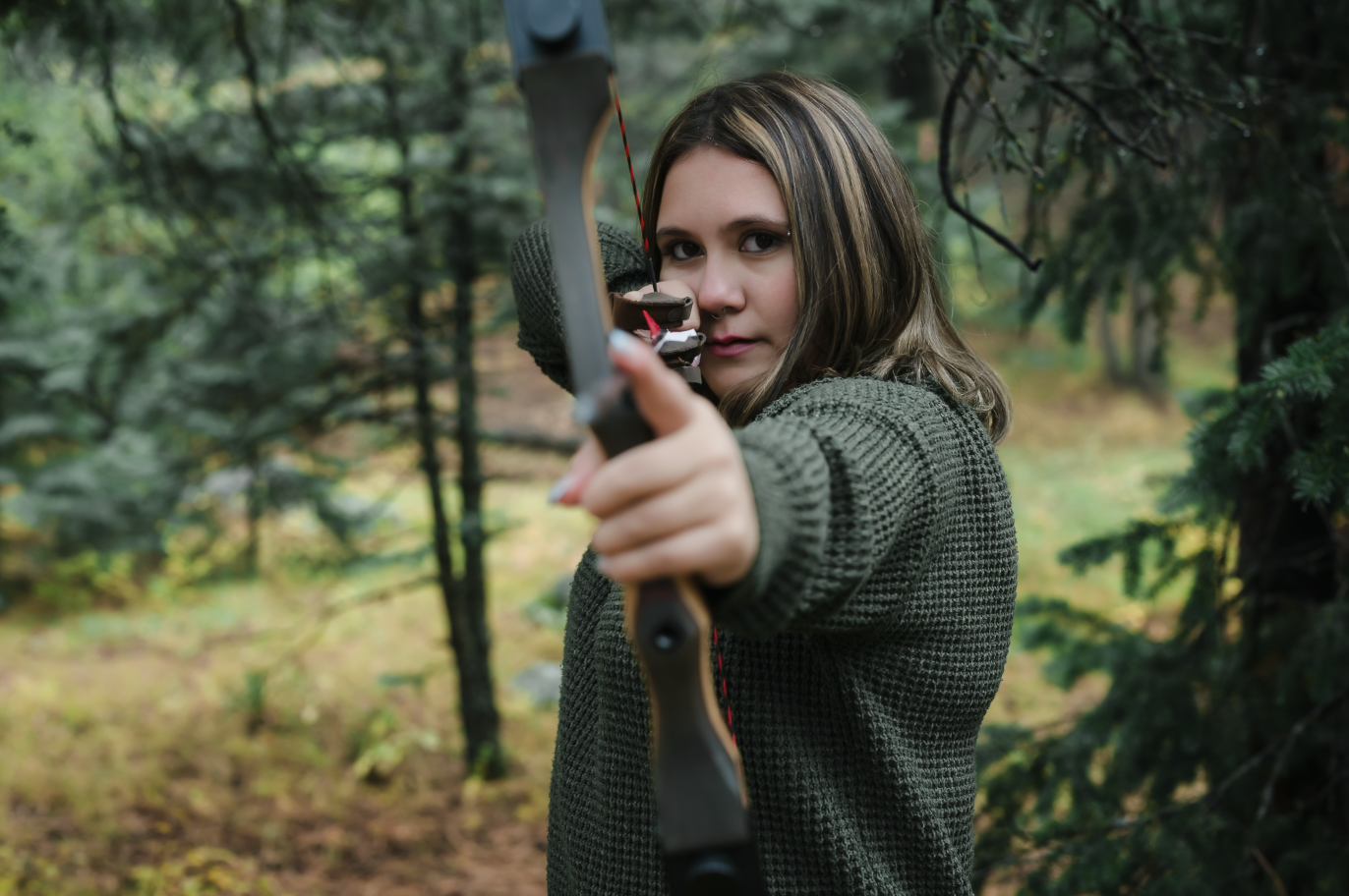 A young woman in outdoor clothing aiming a bow and arrow in a wooded area.
