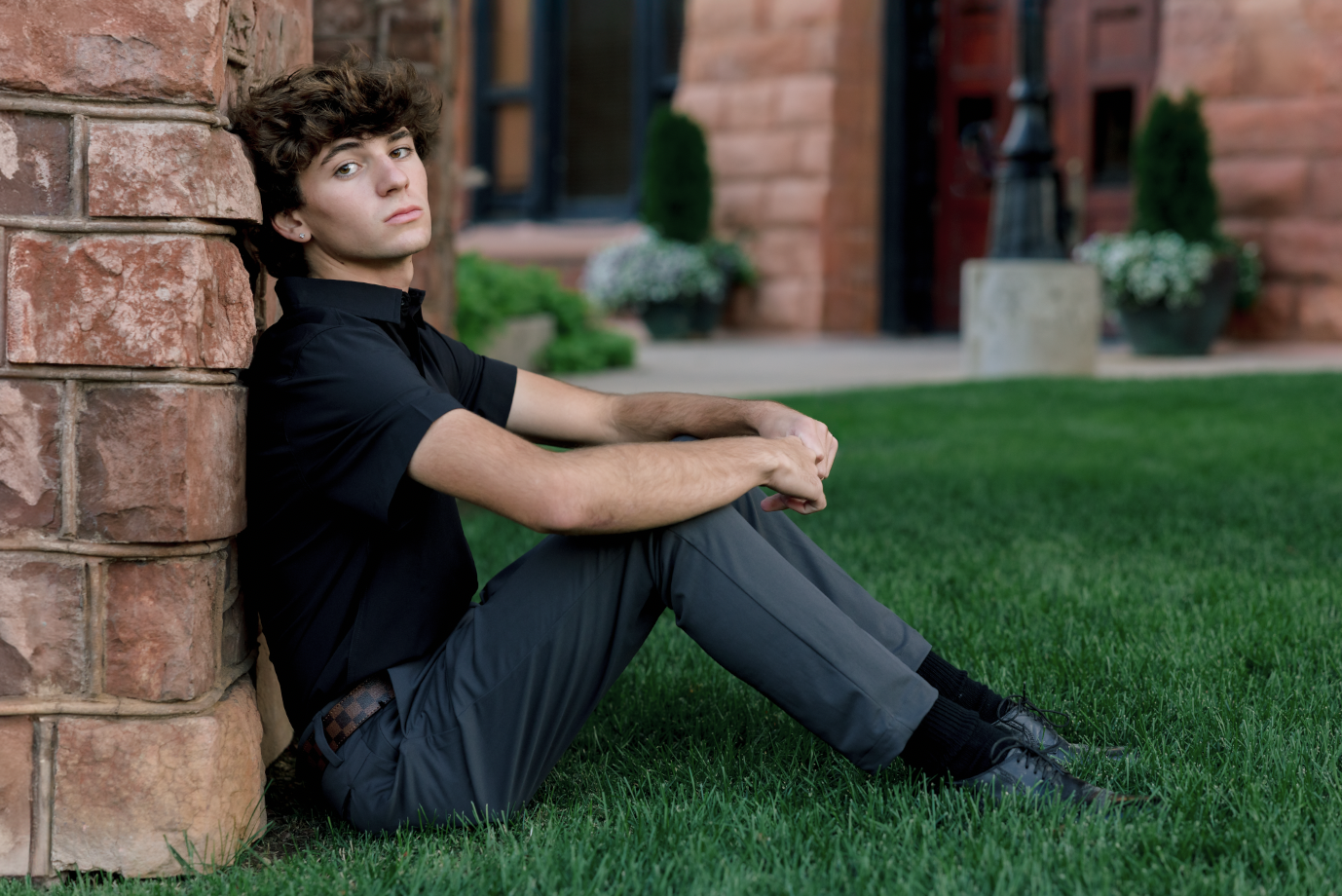 Young man sitting outdoors on grass near a tree, wearing a colorful floral shirt and blue jeans, with a thoughtful expression.