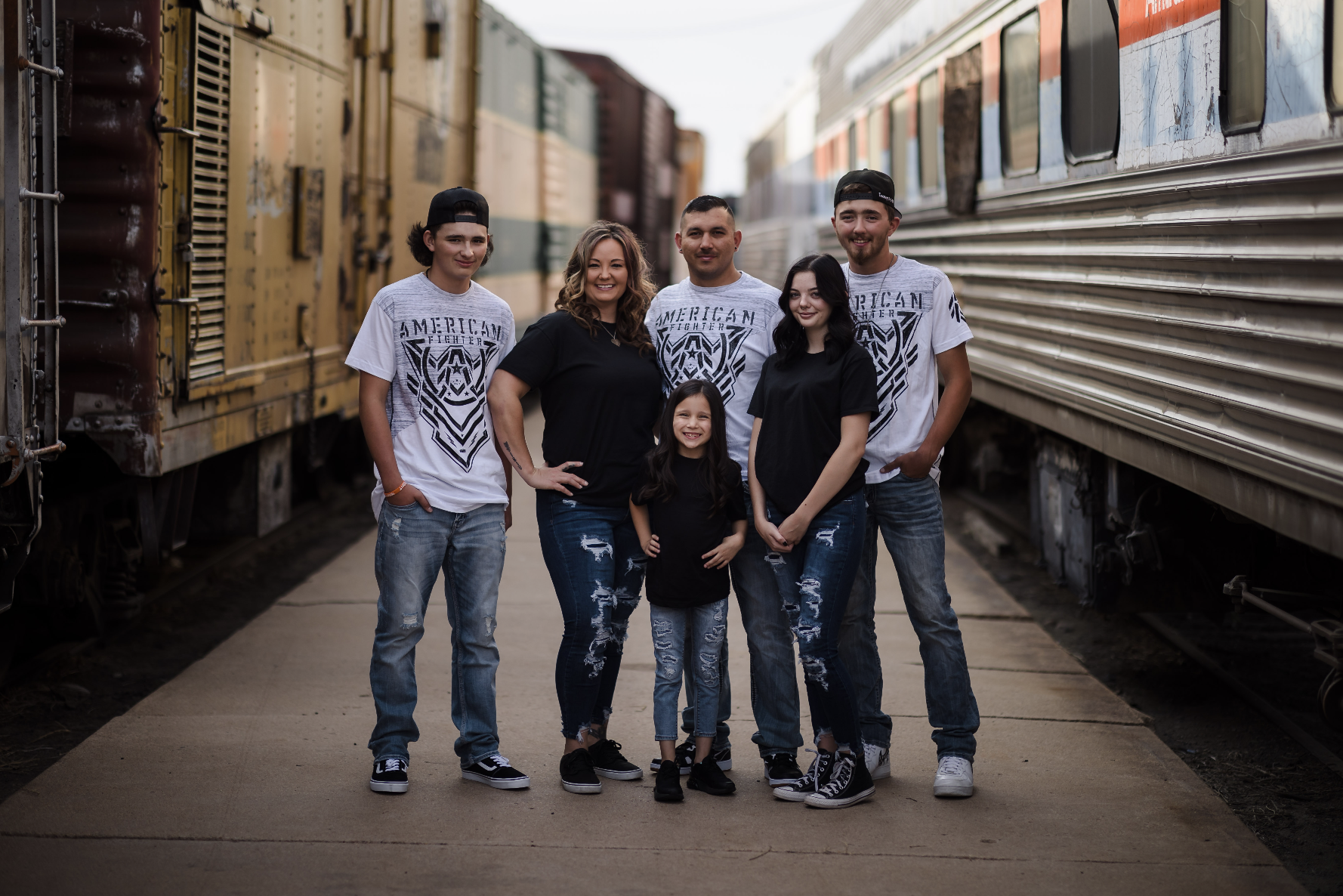 A young man with tousled blond hair and a serious expression, wearing a light gray T-shirt with buttons, standing outdoors in front of a calm body of water and a blurred green landscape.