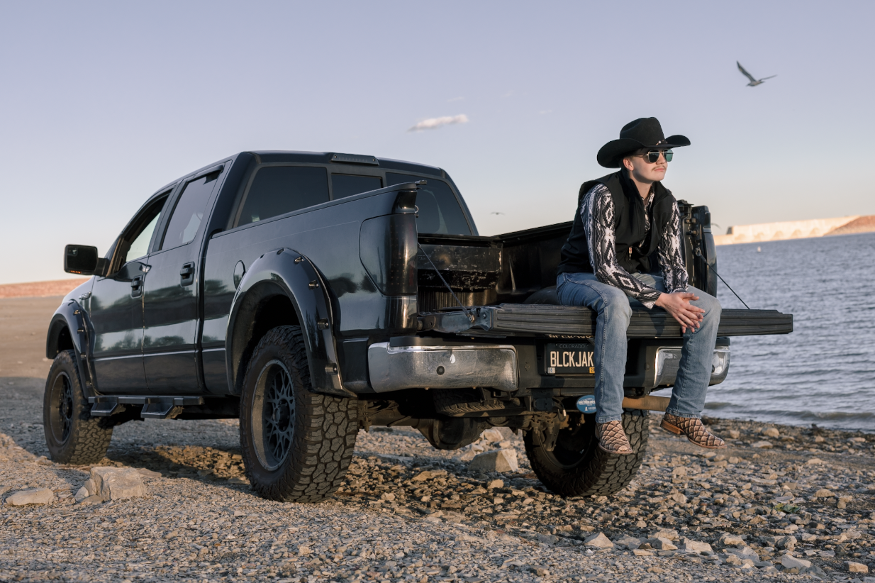 A woman wearing sunglasses, a cowboy hat, and patterned shirt sitting on the tailgate of a black pickup truck parked on a rocky beach near water, with a bird flying overhead.
