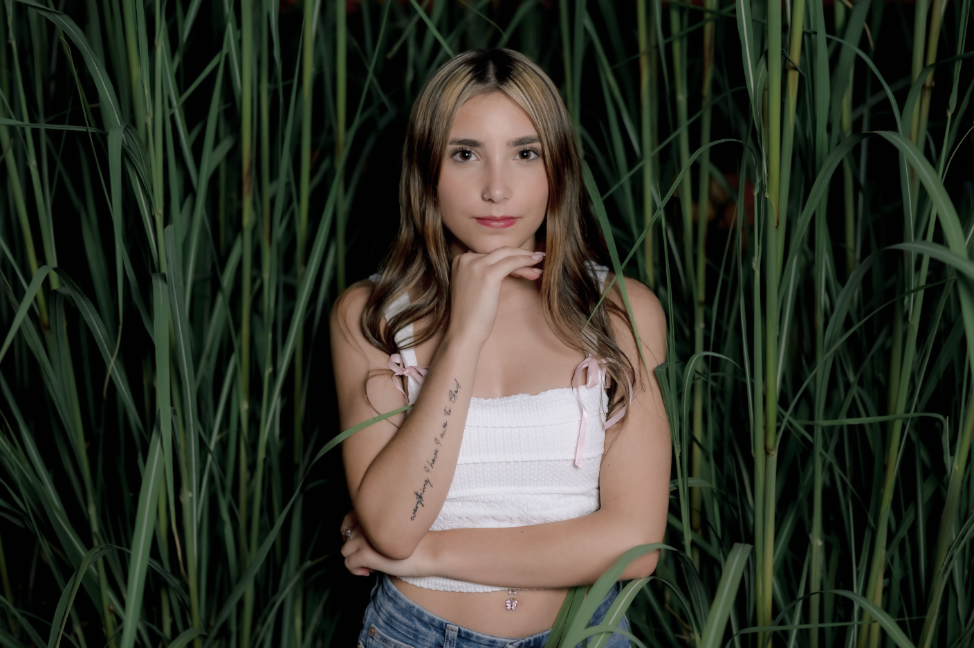A young woman with long, wavy hair standing amidst tall green grass, wearing a white sleeveless top with pink ribbon straps and blue jeans, looking directly at the camera with her hand resting on her chin.
