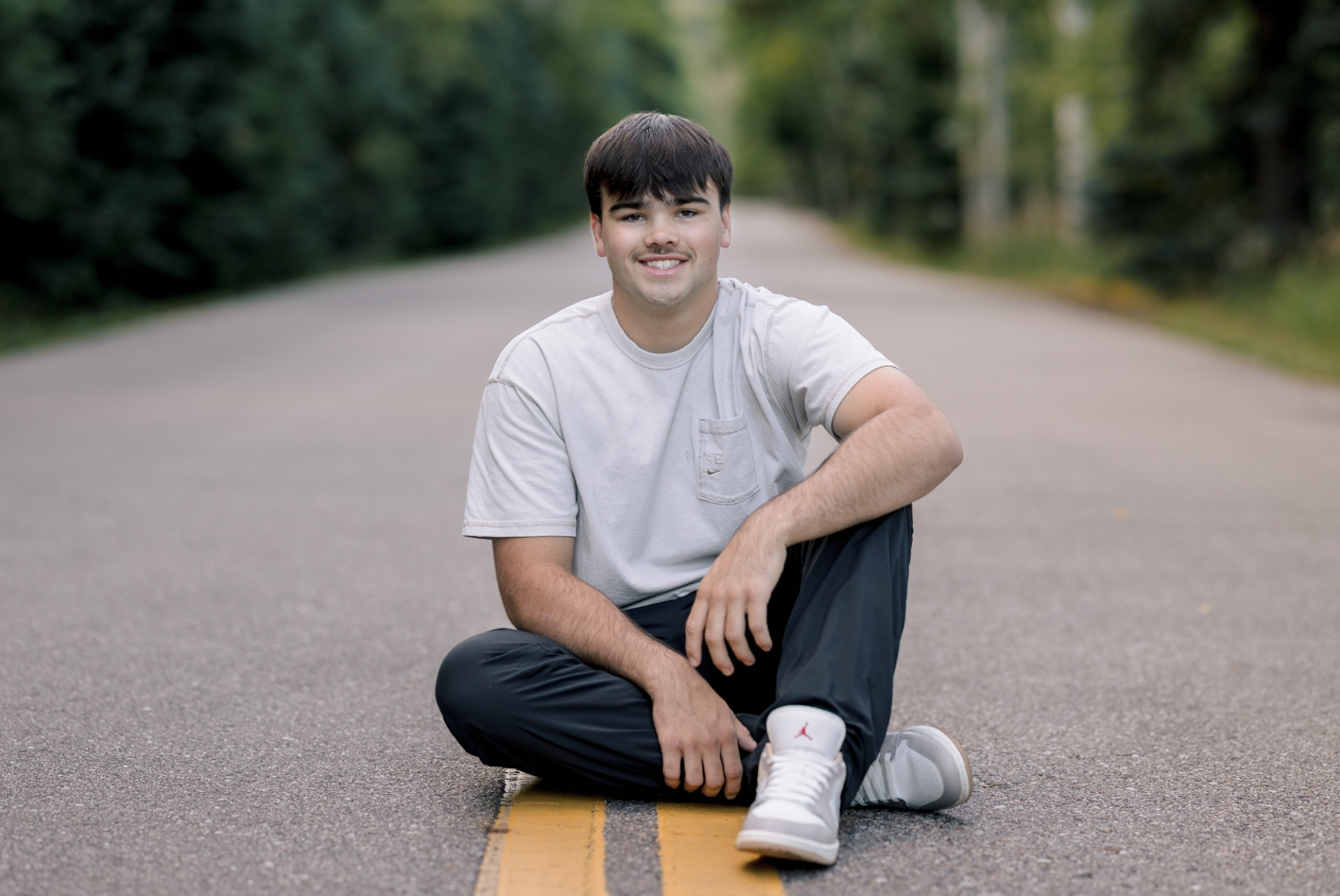 Young man sitting on an empty road with trees in the background, smiling and wearing a gray t-shirt, black pants, and white sneakers.