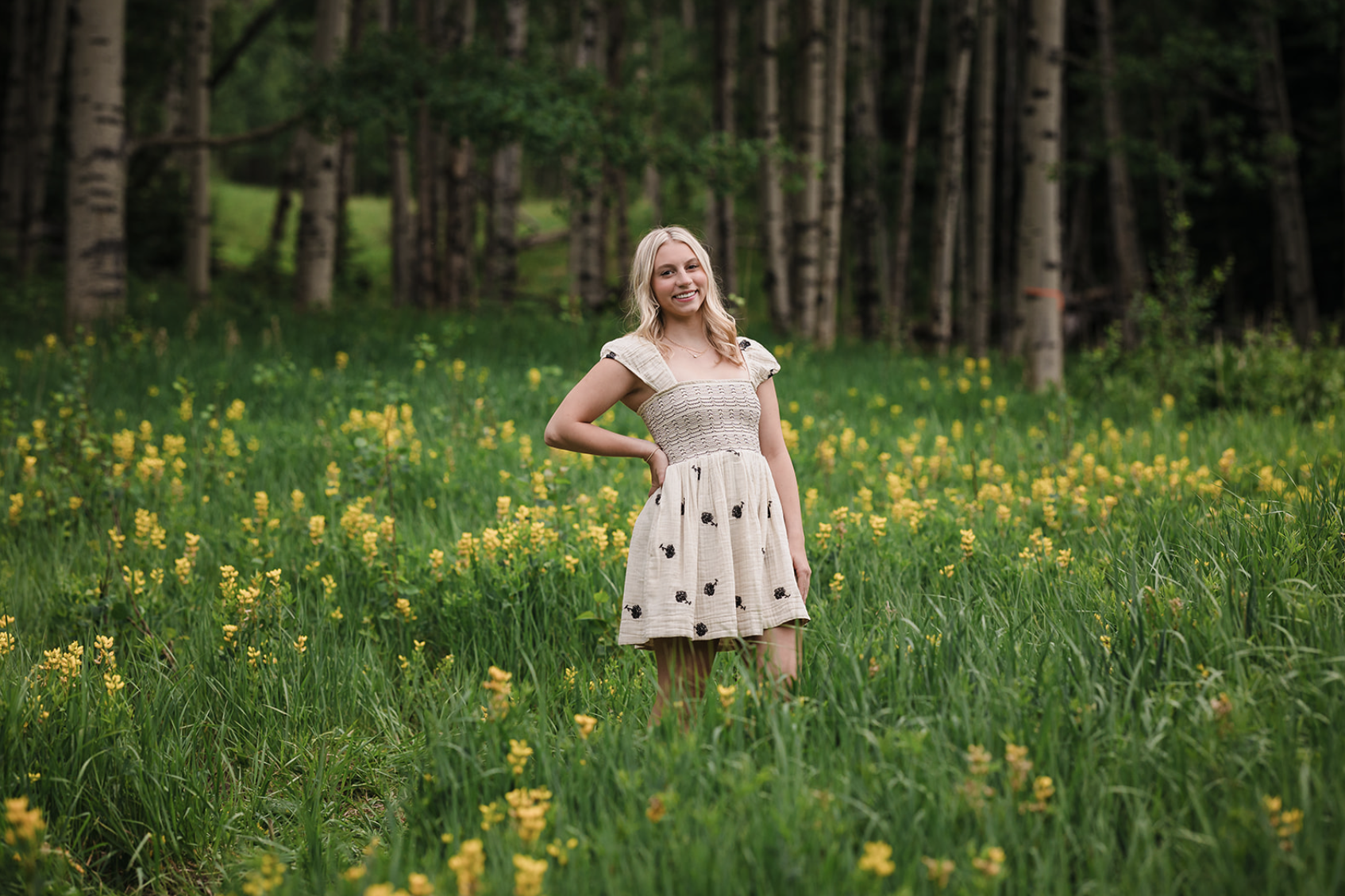 A young woman in a beige dress with black floral patterns smiling in a green field of tall grass and yellow flowers, with a dense forest of tall trees in the background.