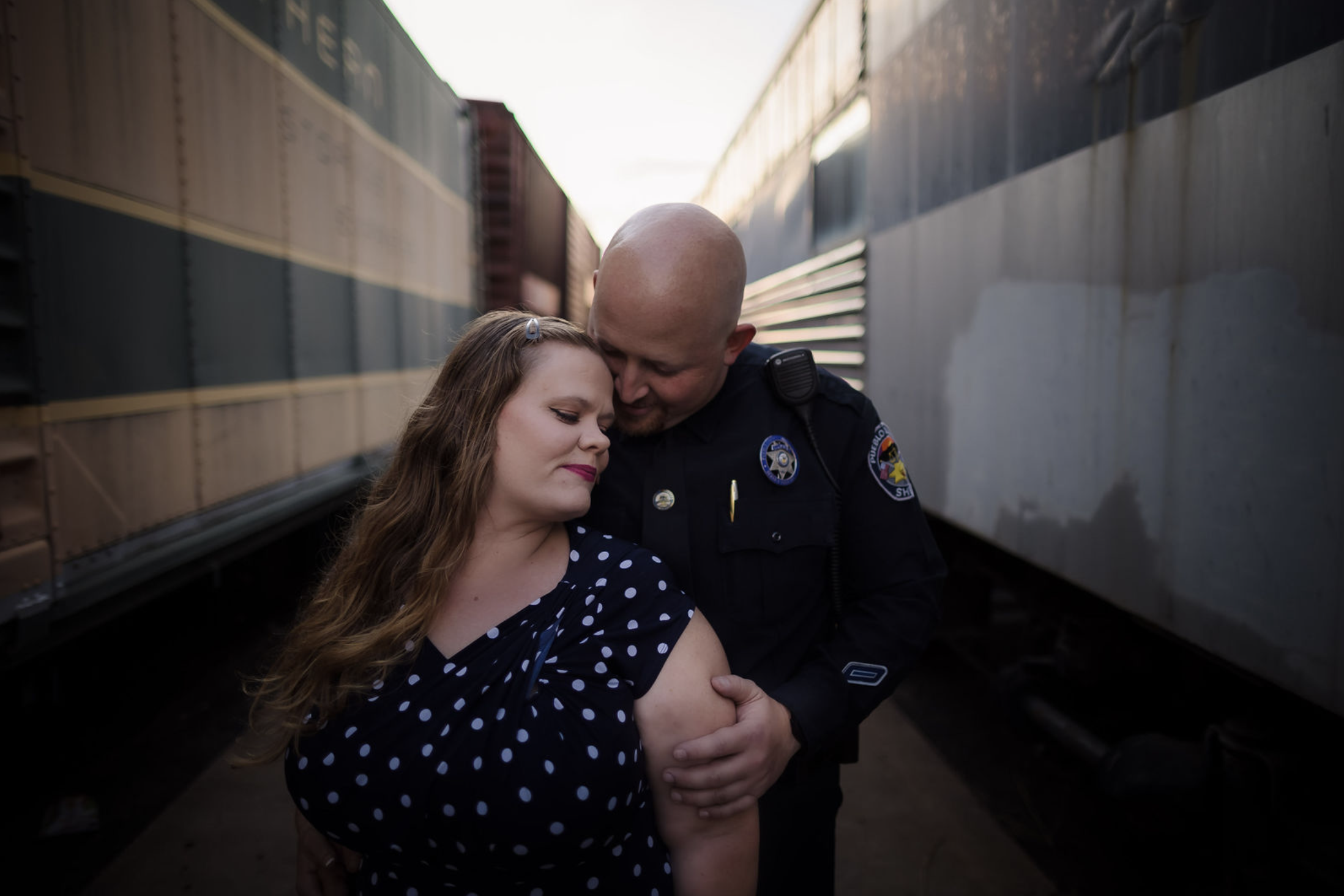 A young girl in a yellow dress with ruffled sleeves and black buttons sitting on a person's shoulder outdoors, smiling and looking at someone out of frame, with trees and greenery in the background.