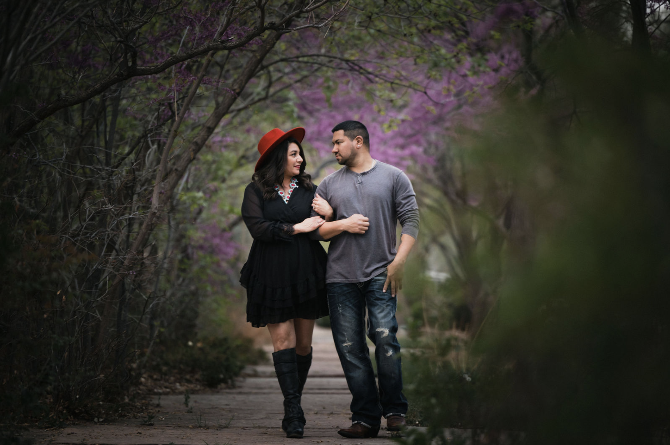 Black and white photo of a man and woman standing outdoors among trees, facing opposite directions with serious expressions, the woman in the foreground and the man slightly behind.