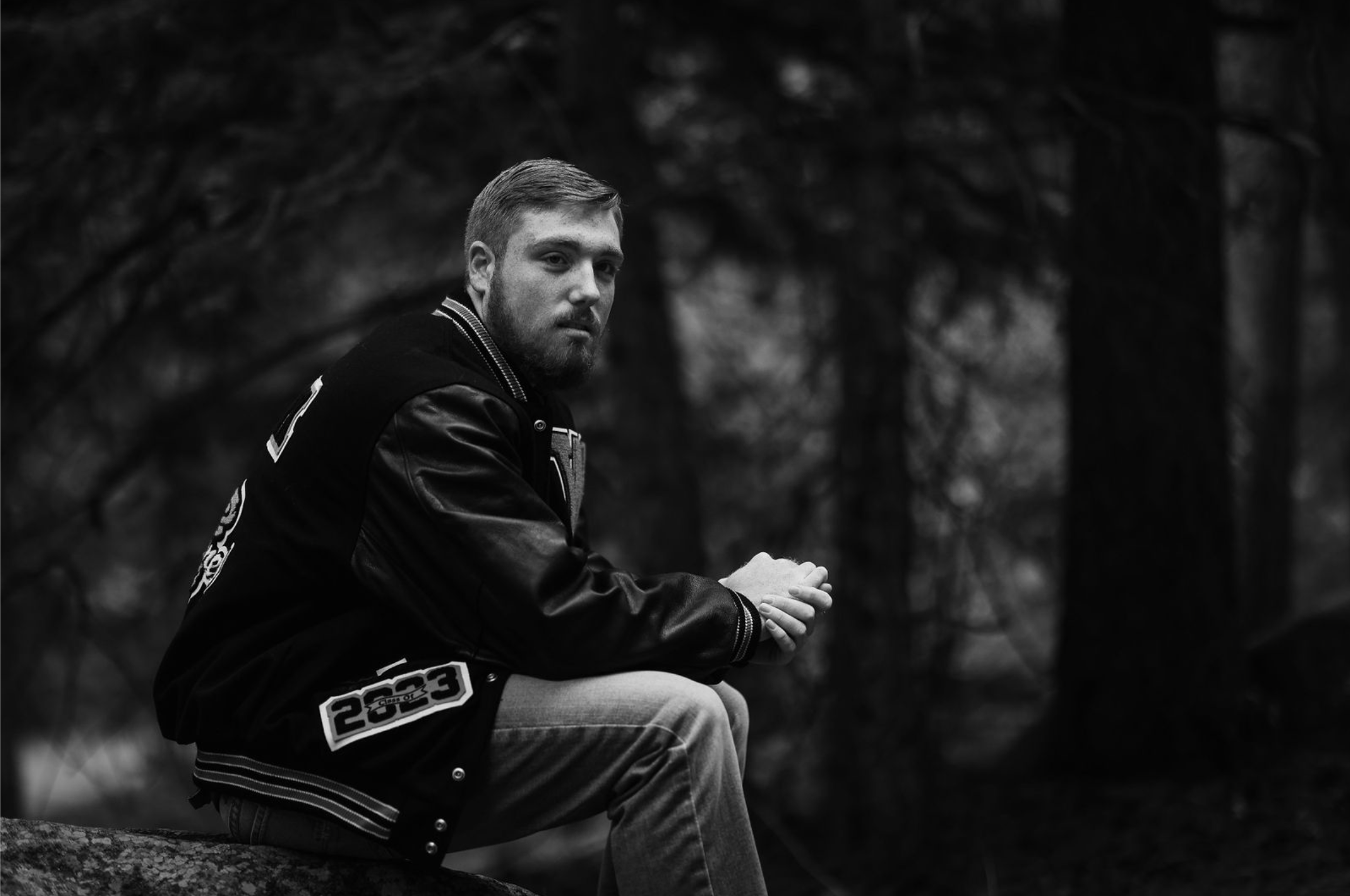 A young man with a beard sitting on a log in a forest, wearing a varsity-style jacket with patches and jeans, looking at the camera.
