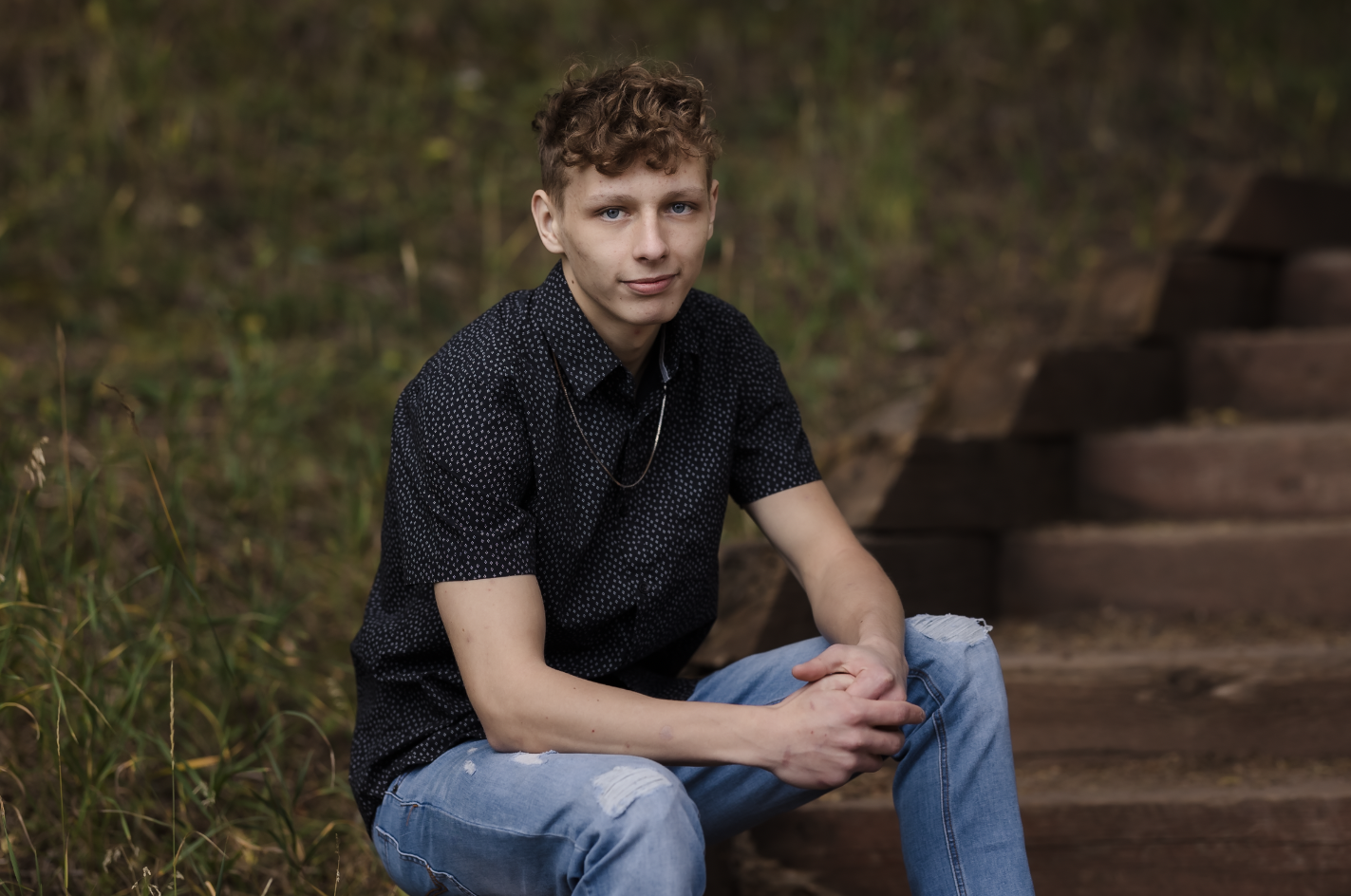 A young man with short curly hair and fair skin sitting on wooden stairs outdoors, wearing a black shirt with white polka dots and ripped jeans, looking at the camera with a slight smile.