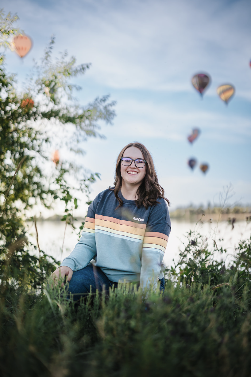 A young woman with glasses and brown hair smiling while sitting outdoors among green grass and plants, with heart-shaped hot air balloons flying in a blue sky in the background.