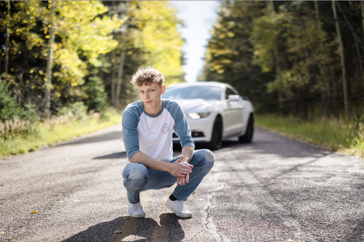 Young man crouching on the road with a white car behind him in a wooded area with yellow leaves.