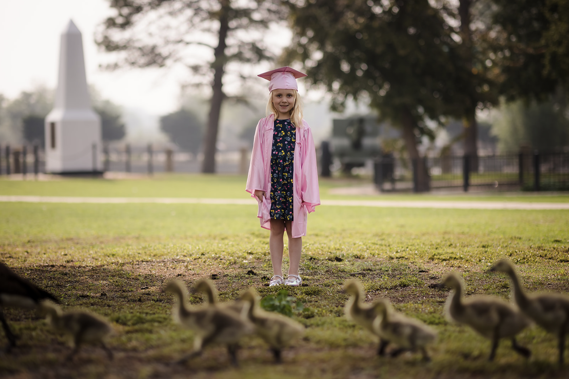 A young girl in a pink graduation cap and gown with a white stole that has colorful letters spelling "Graduated". She is smiling and standing outdoors near a black metal fence, with green trees in the background.