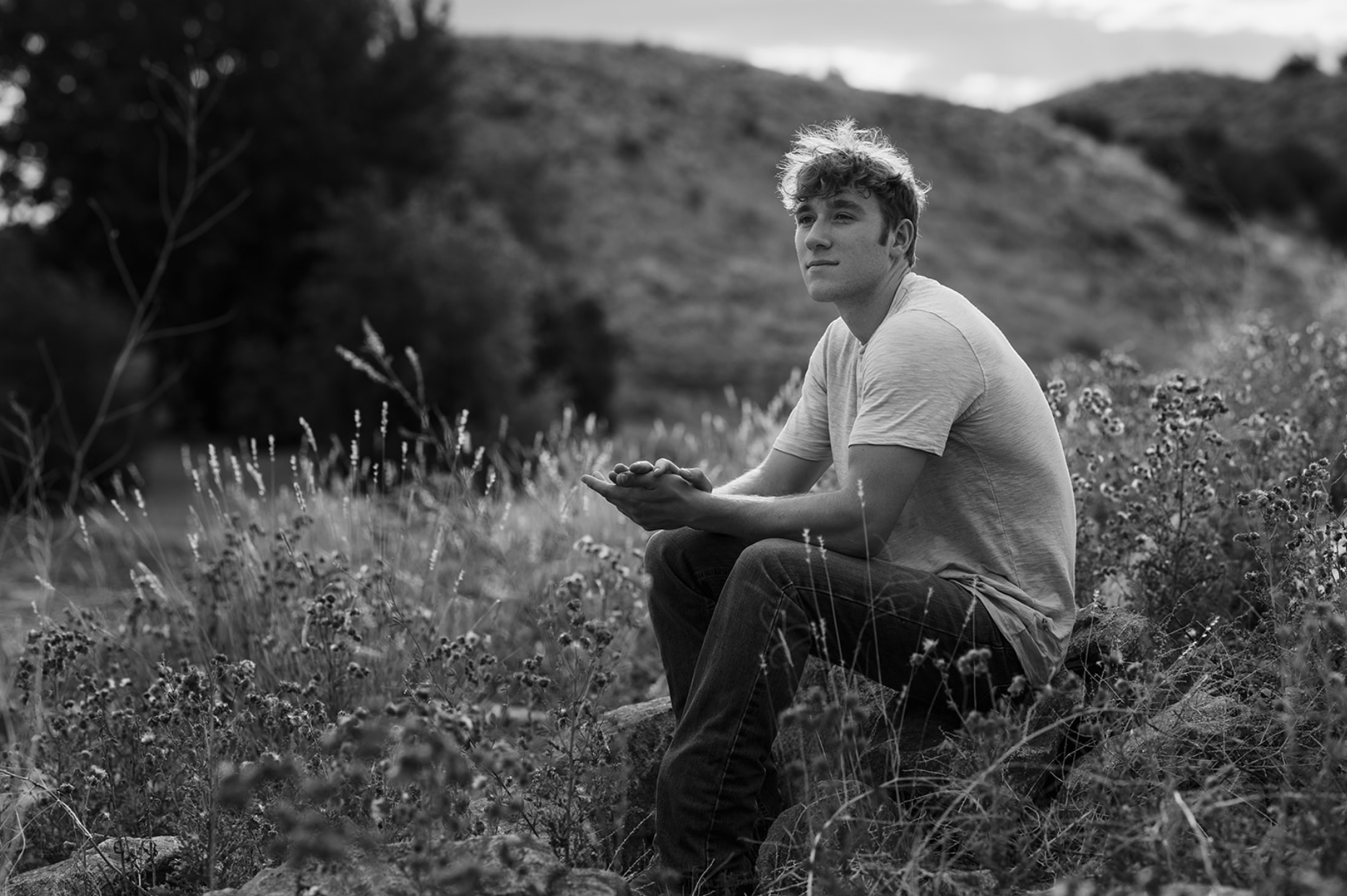 A young woman with dark brown wavy hair and blue eyes smiling outdoors in a park during daytime, wearing a white sweater and layered gold necklaces.