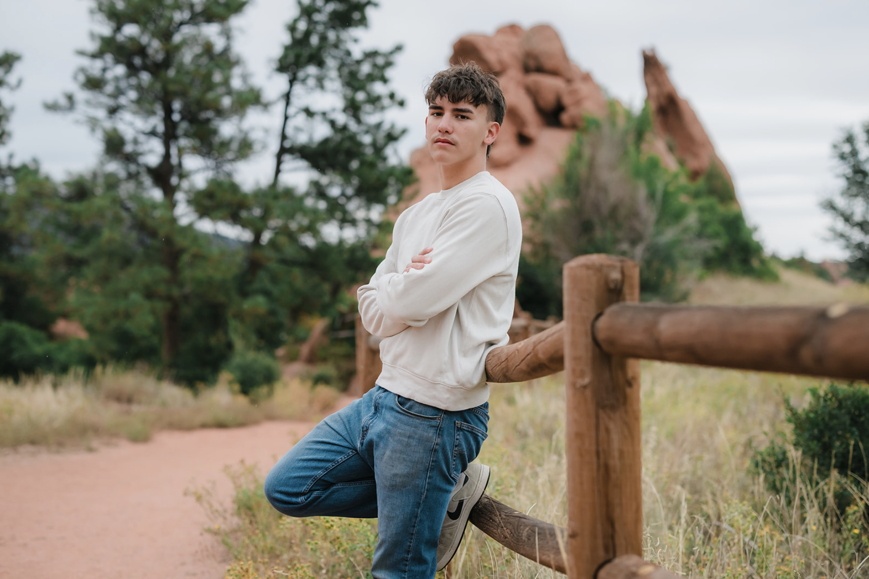 A young man with dark hair, wearing a white sweatshirt, blue jeans, and white sneakers, leaning against a wooden fence on a trail in a natural setting with trees and a large reddish rock formation in the background.
