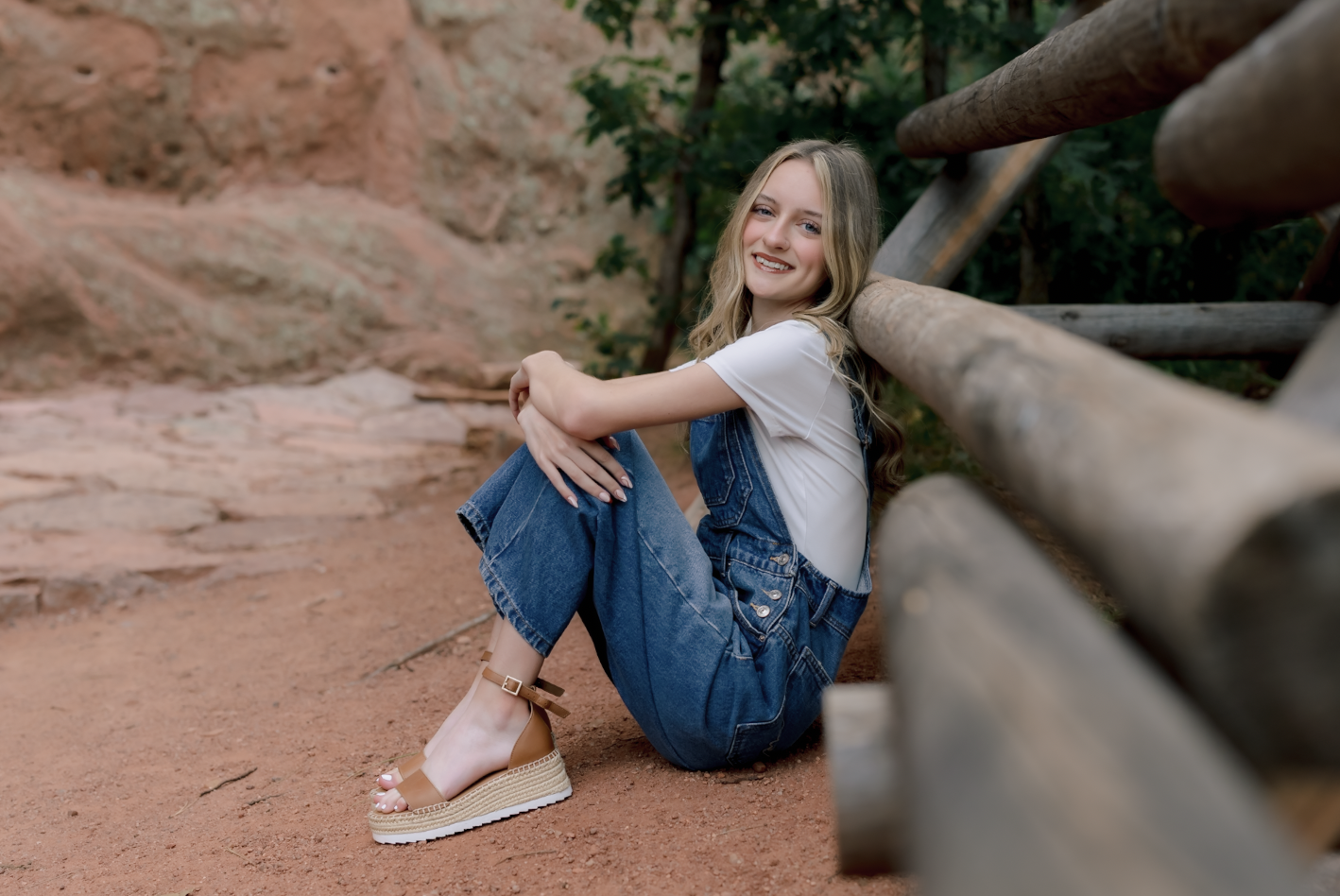 A young woman with long blonde hair, smiling and sitting on a dirt path with reddish soil, leaning against a wooden railing, wearing a white t-shirt, denim overalls, and tan platform sandals, in a natural outdoor setting with rocks and green foliage in the background.