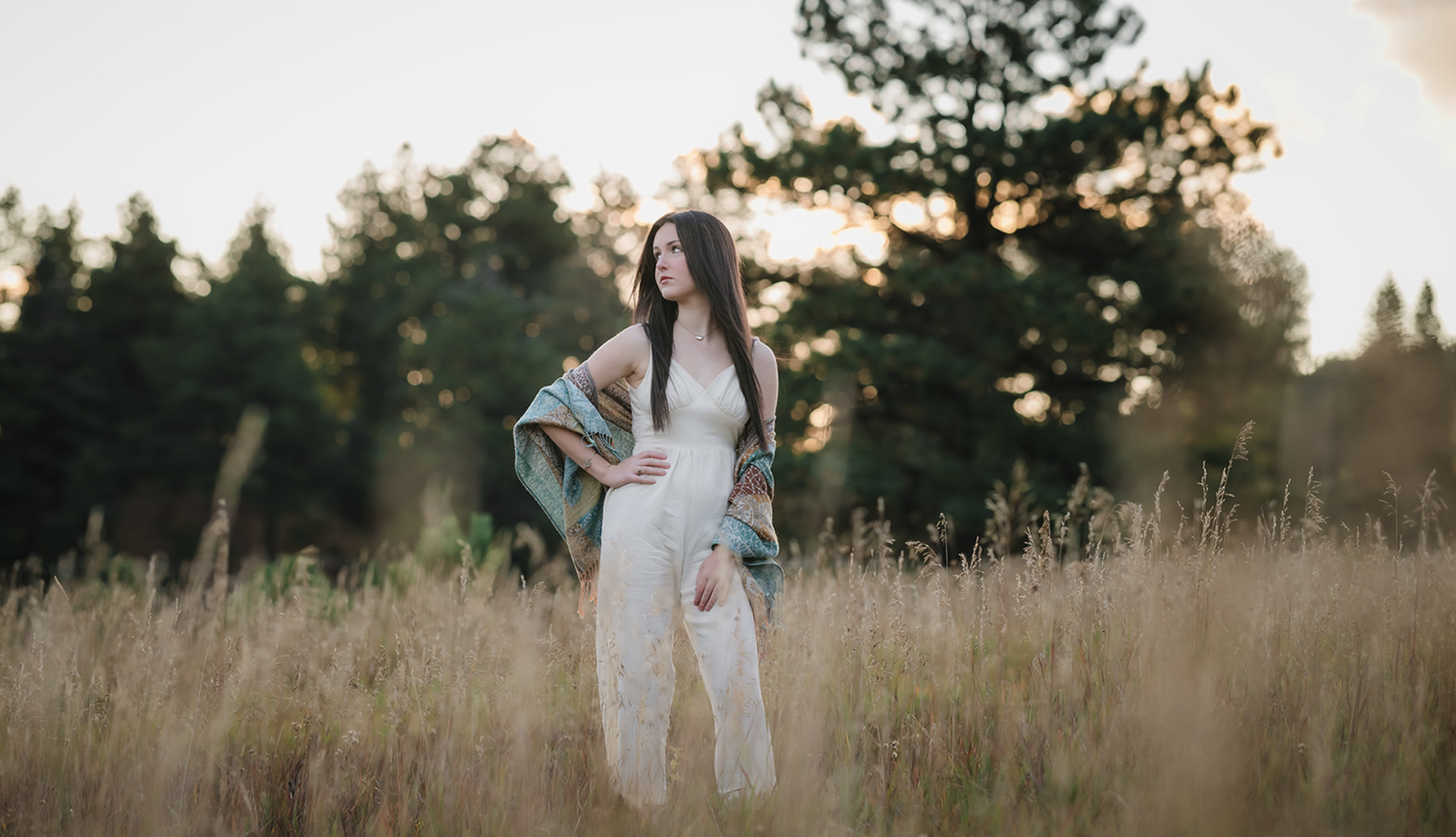 A woman standing in a grassy field during sunset, wearing a white jumpsuit with a colorful shawl draped over her shoulders.