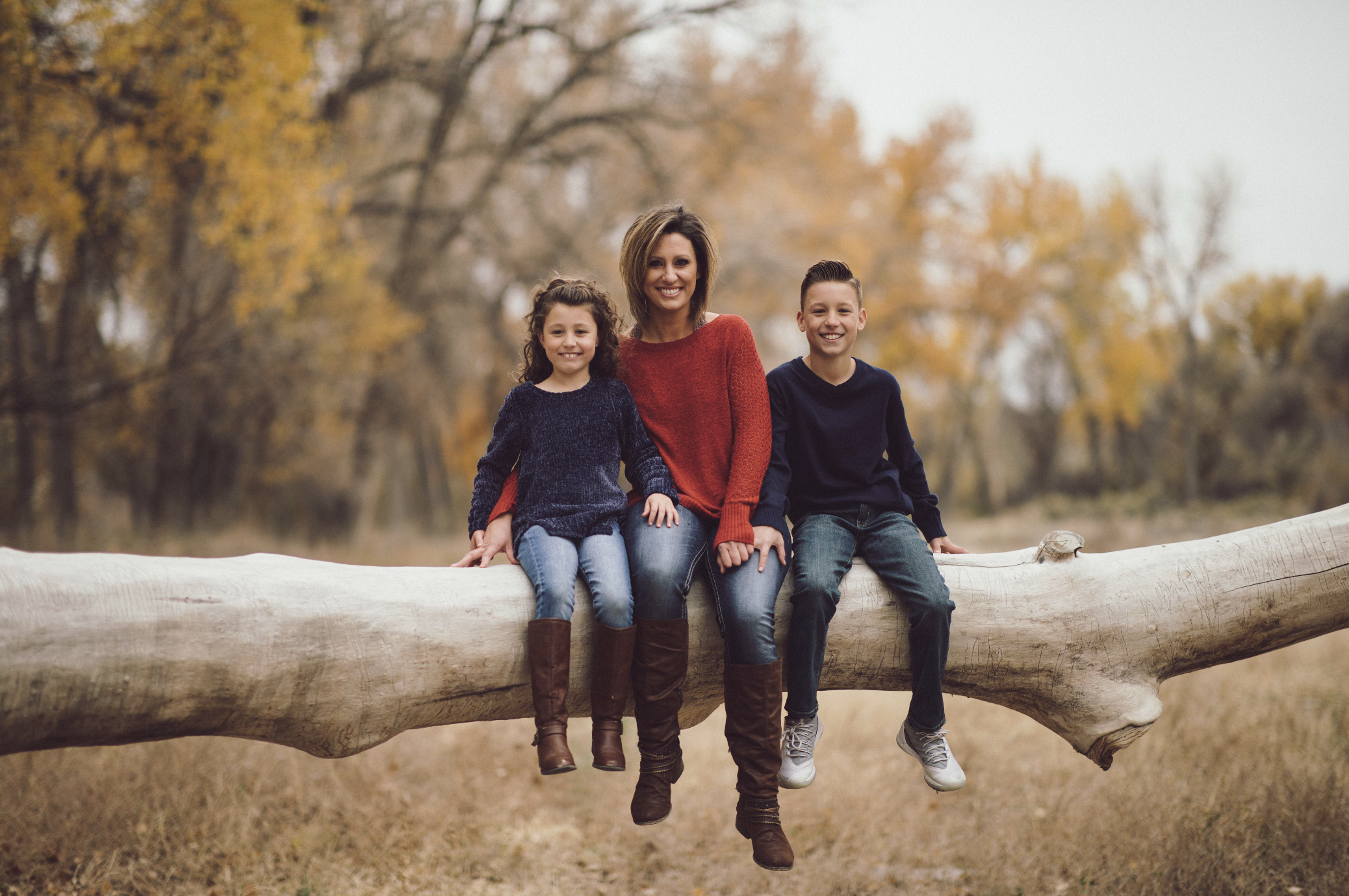 A mature couple stands close to each other by a lake with autumn trees in the background. The woman has gray hair and is wearing a brown sweater with hoop earrings and a necklace. The man has gray hair, glasses, and is wearing a plaid shirt. They are holding hands and looking at the camera.