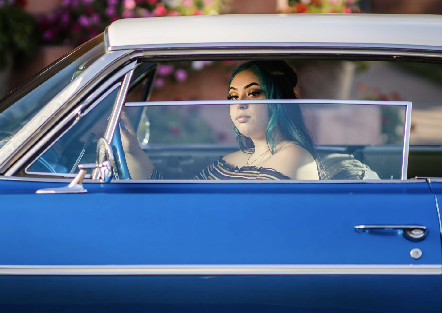 A young woman with teal hair sitting in the driver's seat of a vintage blue car, looking out the window with a serious expression, wearing an off-the-shoulder striped top.