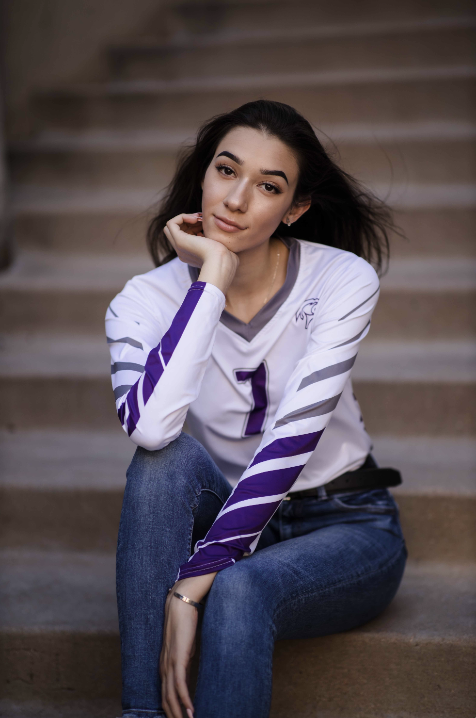 Young woman sitting on steps, resting her chin on her hand, wearing a white sports jersey and jeans.