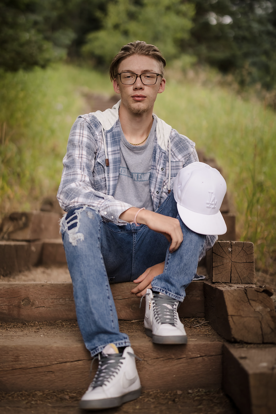 Young man sitting on wooden steps outdoors, wearing glasses, a gray t-shirt, a plaid shirt, ripped jeans, white sneakers, and holding a white baseball cap.