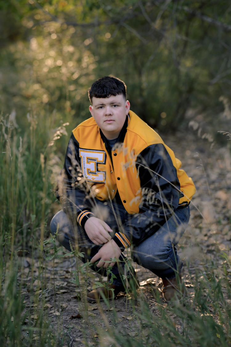 A young man crouching outdoors in a wooded area with green trees and plants, wearing a yellow and black varsity jacket with a letter "E" on the front.