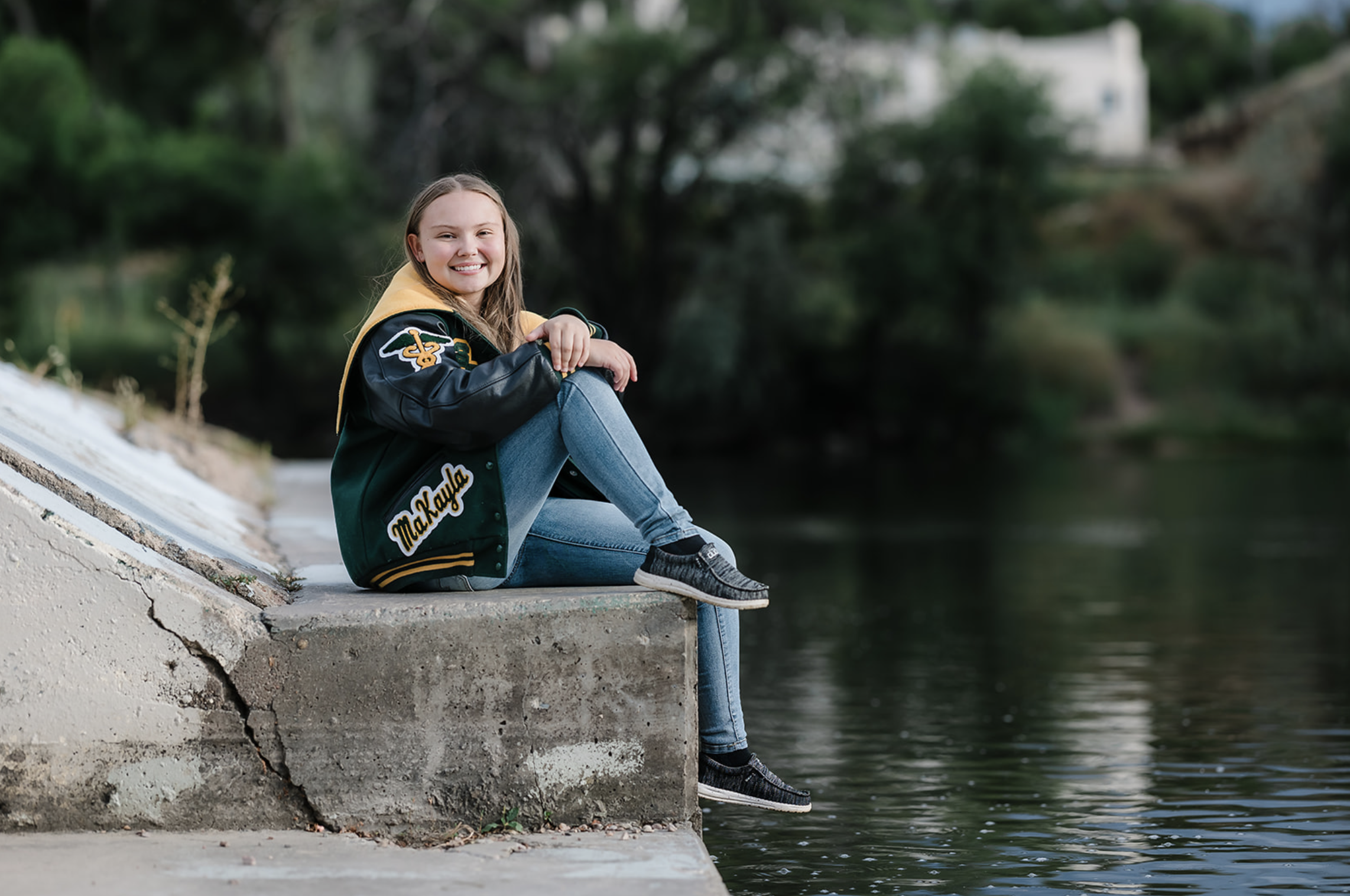 A young woman with long hair sitting on a concrete ledge by a river, smiling, wearing a black and yellow letterman jacket, blue jeans, and black sneakers, with trees and houses in the background.