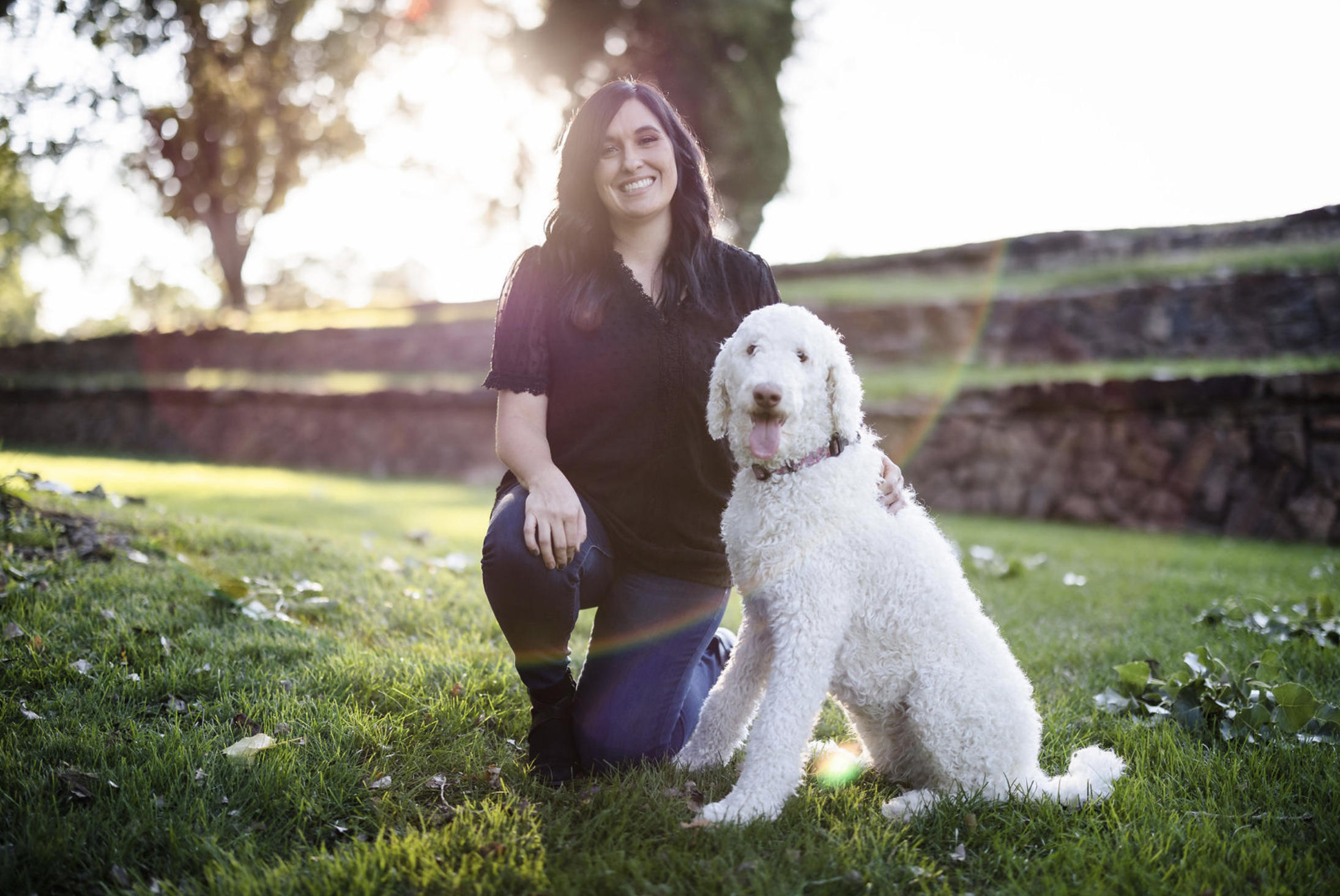 A pregnant woman in a yellow dress standing outdoors in a park at sunset, smiling and holding her belly, with trees and grass in the background.