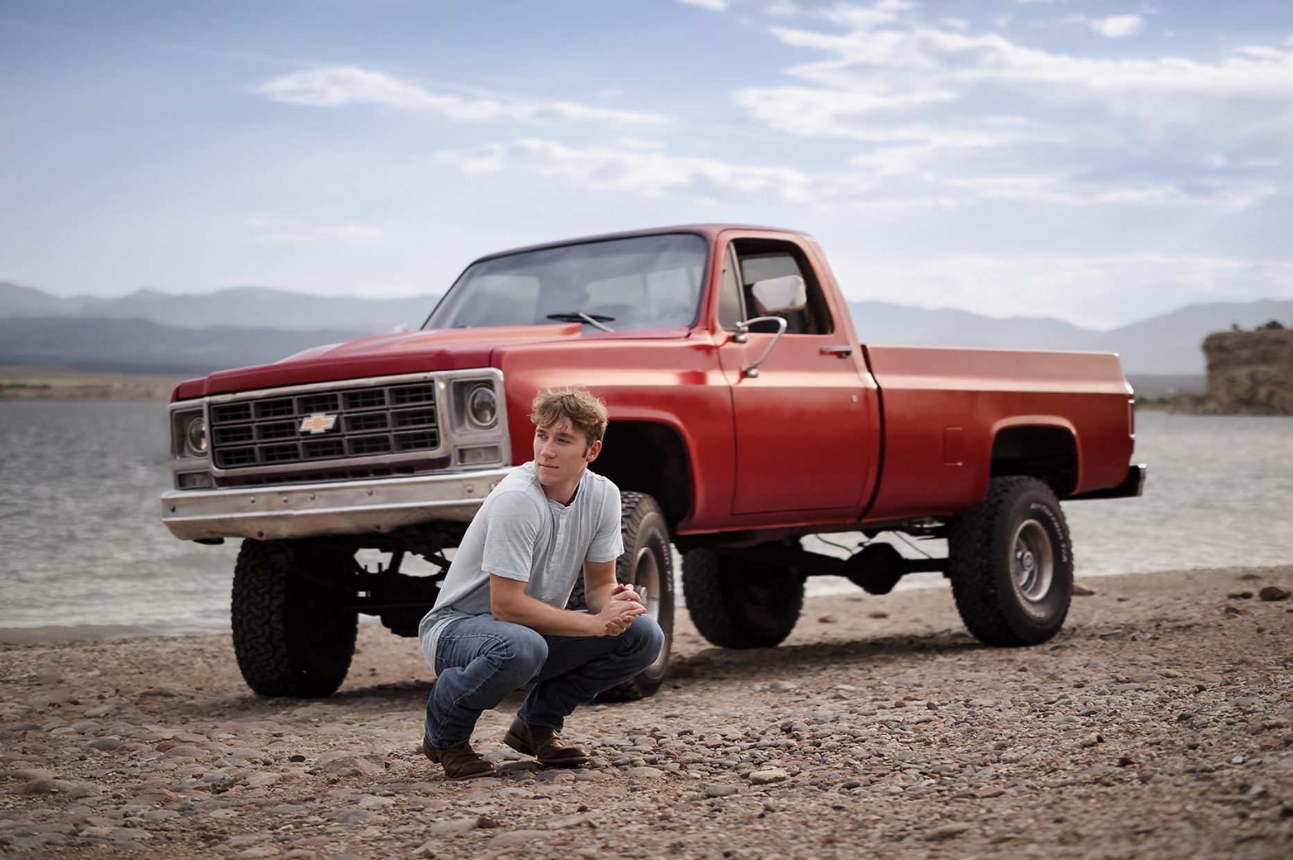 Young man crouching on a rocky shore next to a red vintage pickup truck with a Chevrolet logo, near a body of water with mountains in the background under a partly cloudy sky.