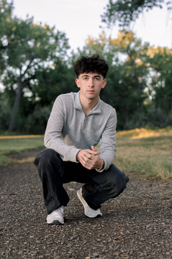 A young man with dark hair and fair skin crouching on a gravel path outdoors, wearing a gray quarter-zip sweater, black pants, and white sneakers. Behind him are green trees with some autumn foliage.