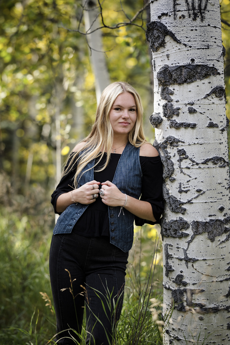 A young woman with long blonde hair standing outdoors next to a white birch tree with black markings. She is wearing a black top with shoulder cut-outs, a denim vest, and black pants, and is holding her hands near her chest, in a forest setting with green foliage in the background.