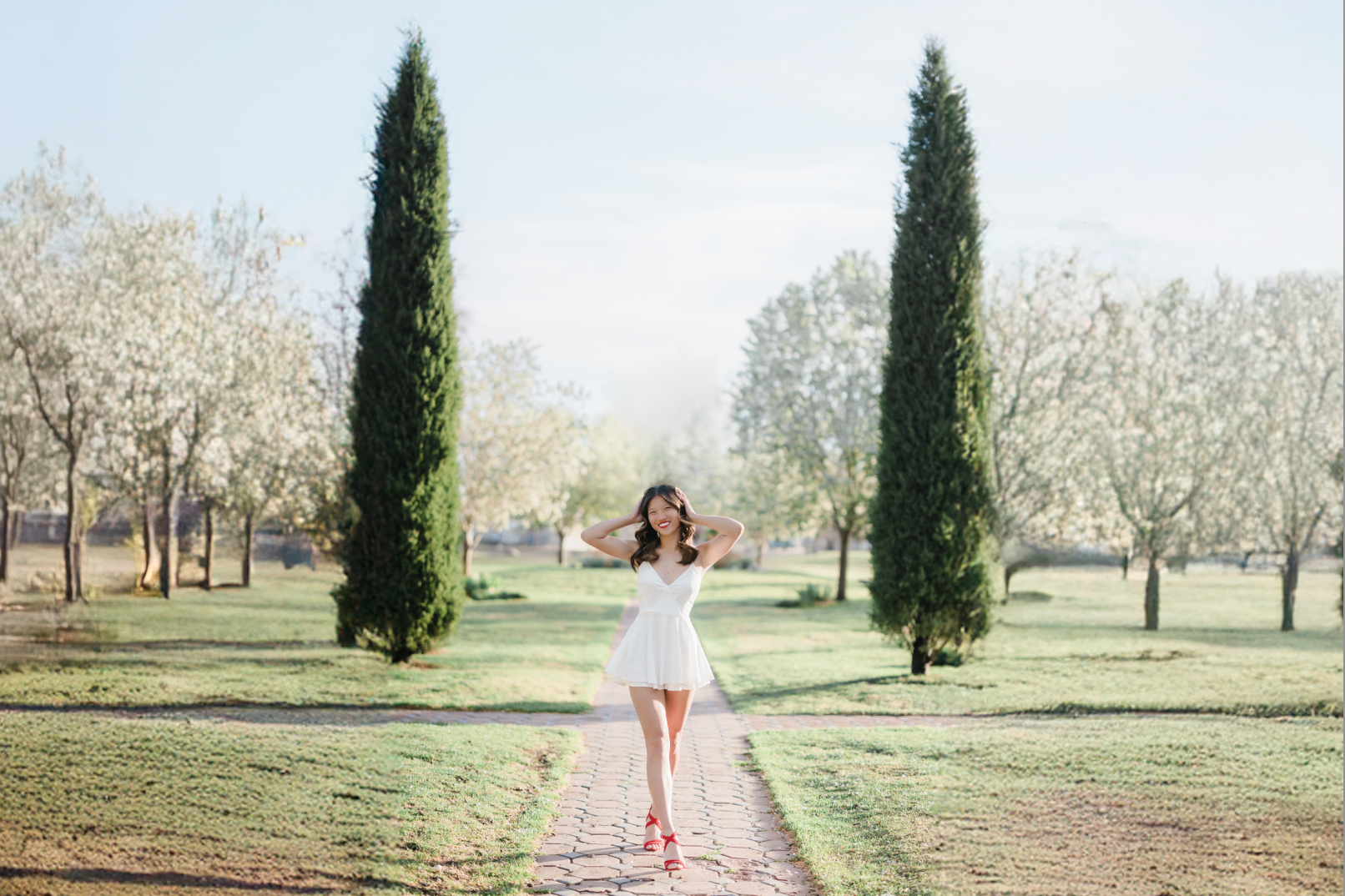 A woman in a white dress and red sandals walking on a brick path in a park with green grass, tall trees, and clear blue sky.
