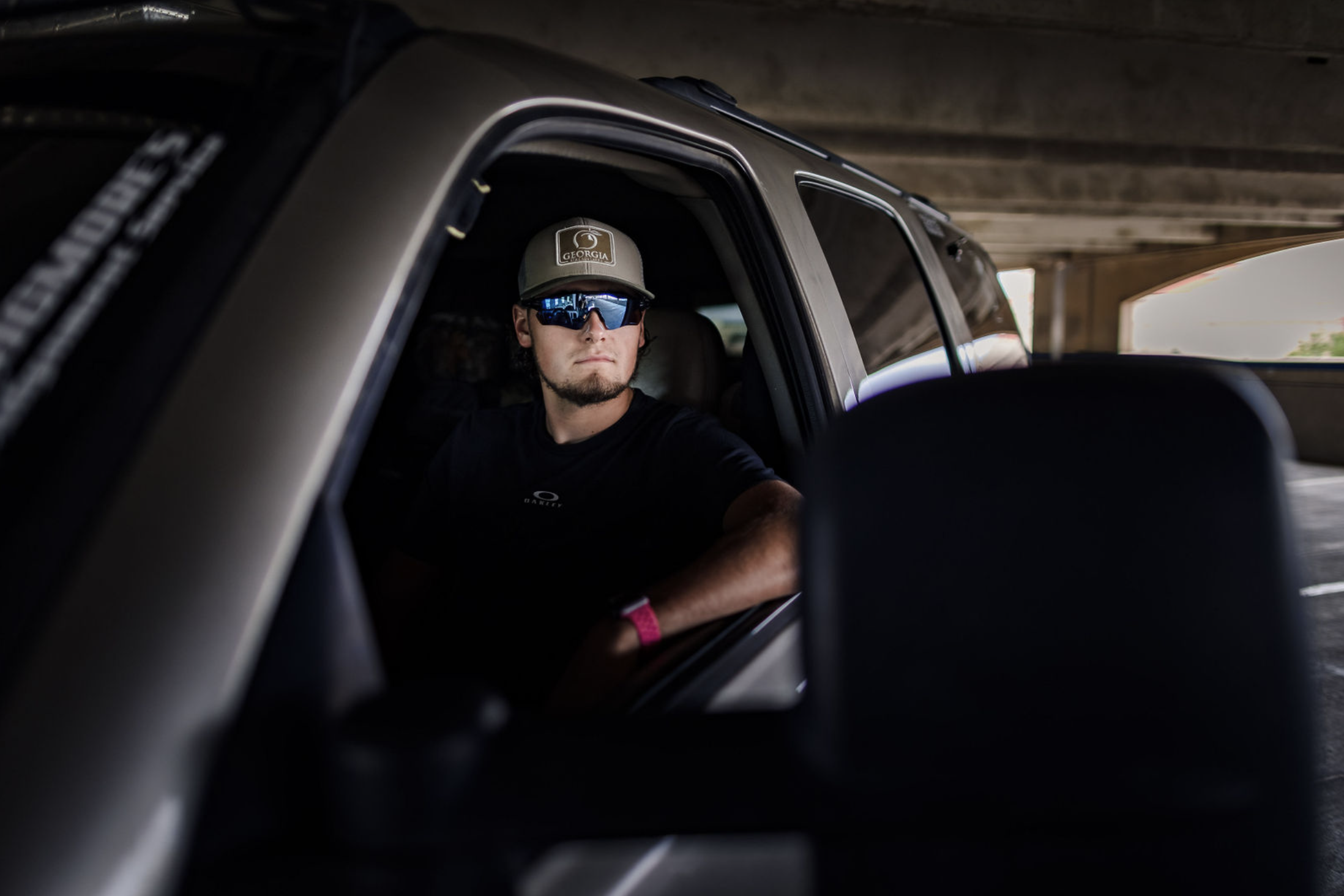 A man wearing sunglasses, a hat labeled 'Georgia,' and a black t-shirt sitting in the driver's seat of a vehicle under a bridge or overpass.