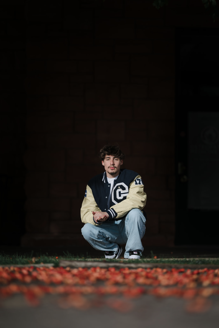 A young man in a varsity jacket crouching on the sidewalk at night with scattered red leaves in the foreground, against a dark brick wall.