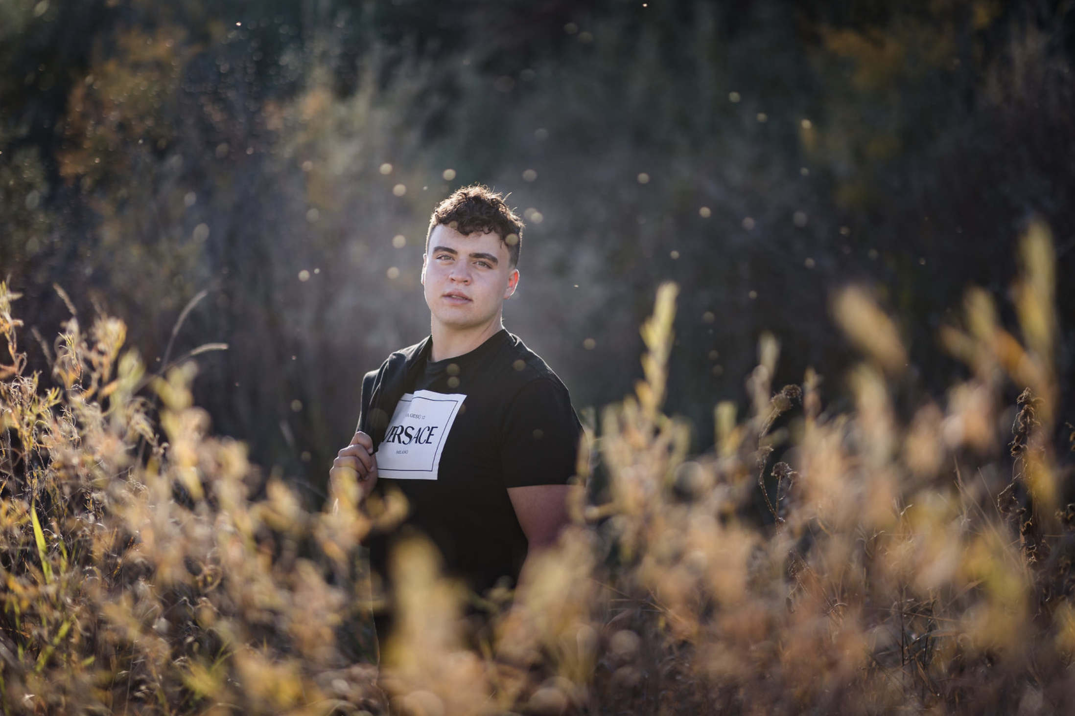 A young man with tousled blond hair and a serious expression, wearing a light gray T-shirt with buttons, standing outdoors in front of a calm body of water and a blurred green landscape.